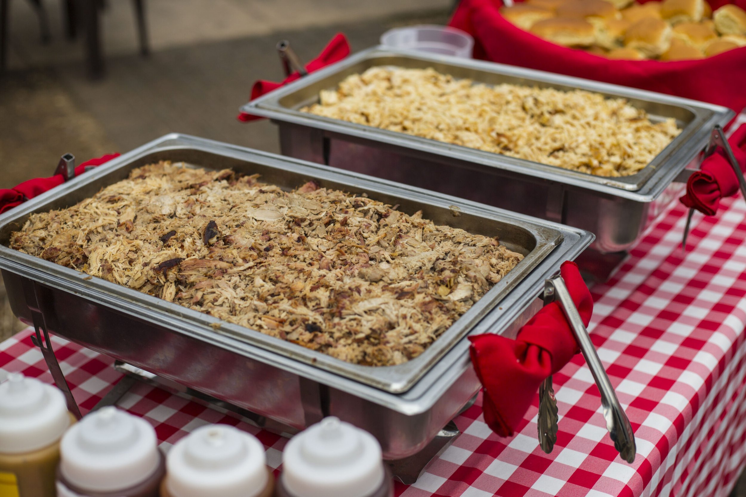 Two large stainless steel chafing dishes filled with shredded meat and rice, placed on a red and white checkered tablecloth, with condiments visible in the foreground.