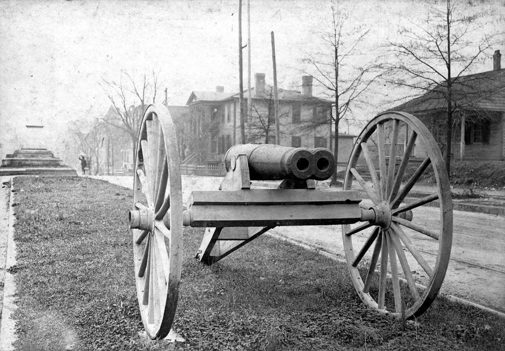 Old black-and-white photo of a cannon on a two-wheeled cart in a park or street, with buildings, trees, and a monument in the background.