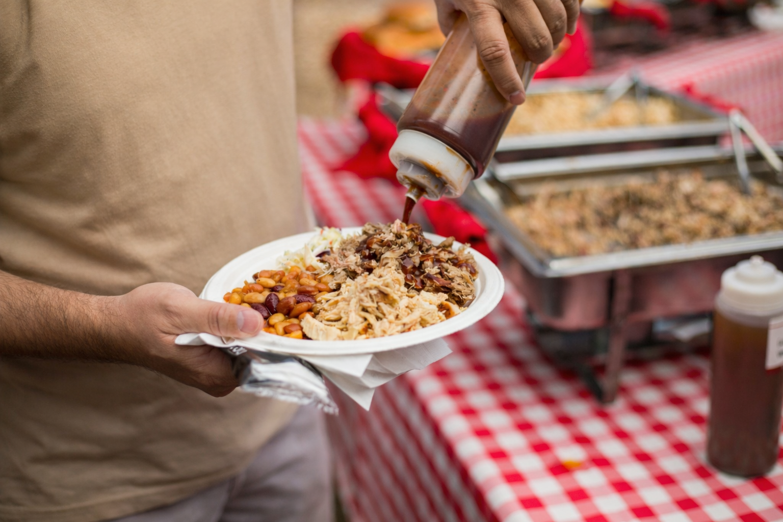 Person holding a paper plate with beans, shredded meat, and coleslaw, while pouring barbecue sauce on the food at a food stand with trays of barbecue in the background.
