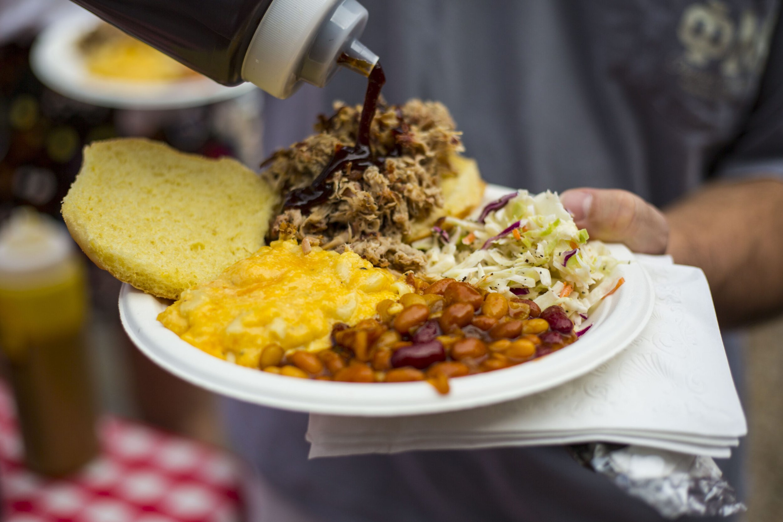 A plate of barbecue with sliced cornbread, scrambled eggs, baked beans, and coleslaw, with barbecue sauce being poured on the meat.