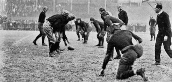 A group of men playing soccer on a field, with some bending down to reach the ball while others watch or stand nearby, in a black and white photo.