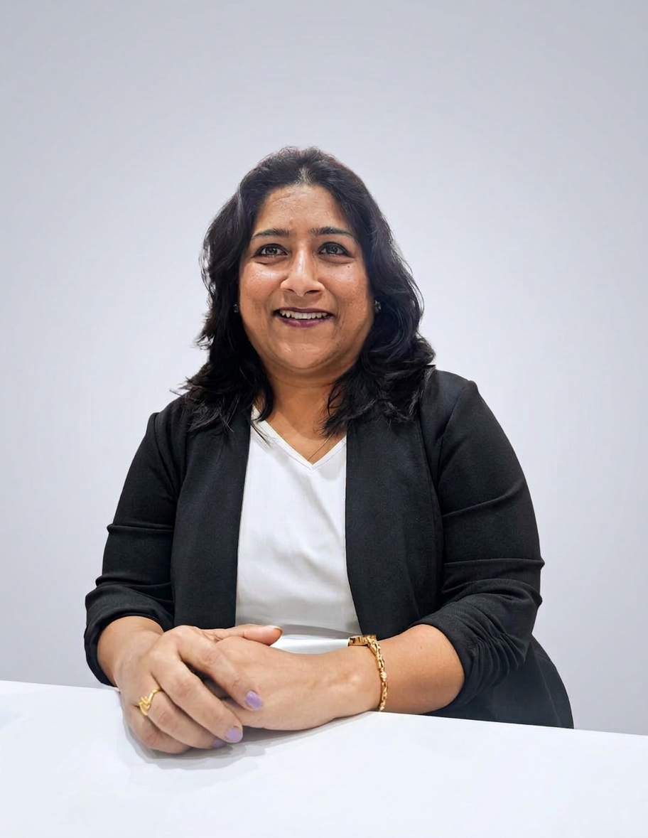 A woman with shoulder-length black hair, wearing a black blazer over a white top, smiling and sitting at a white table against a plain white background.