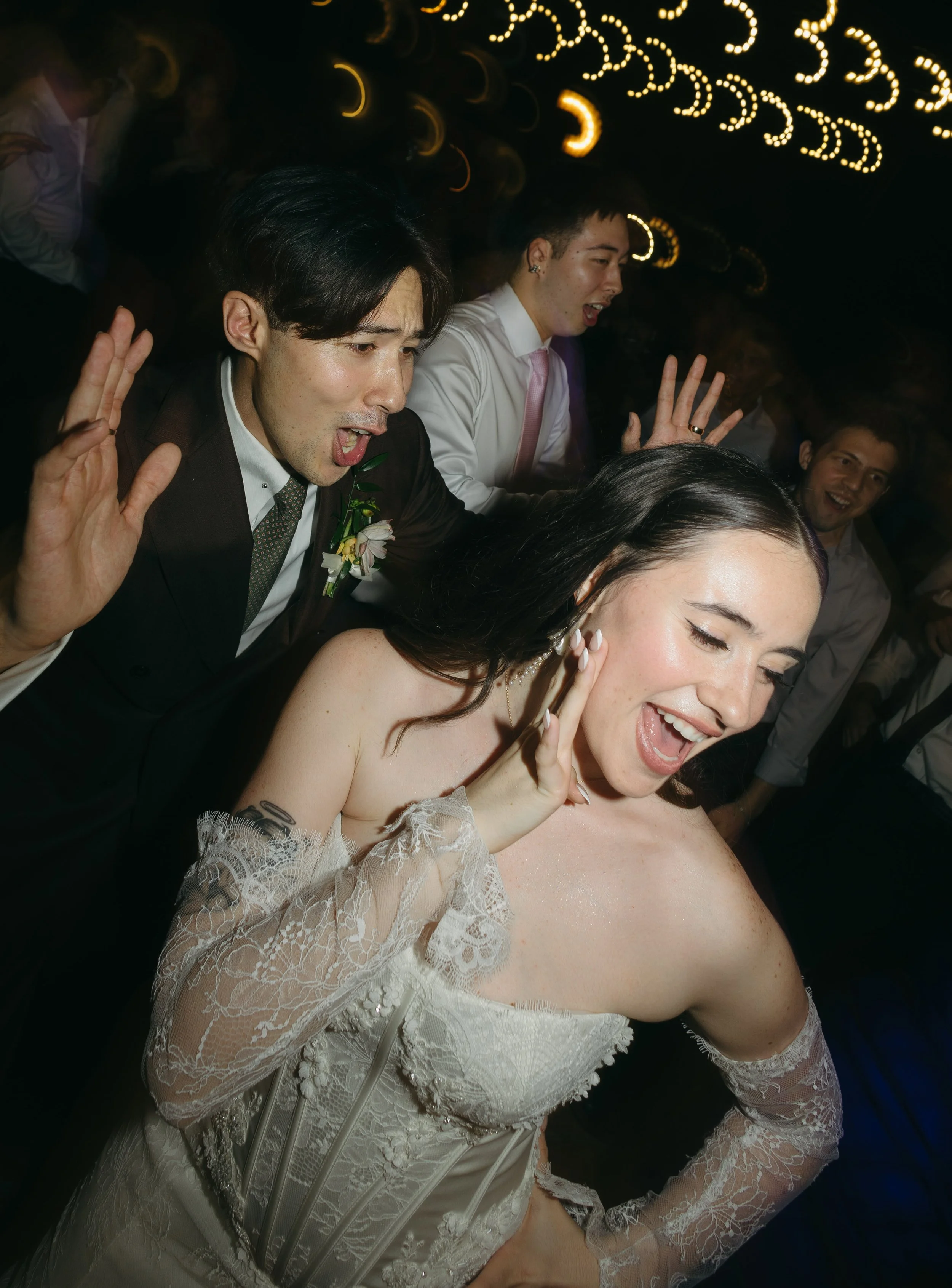 People dancing at a celebration, including a woman in a wedding dress and men in formal suits, under colorful lights.