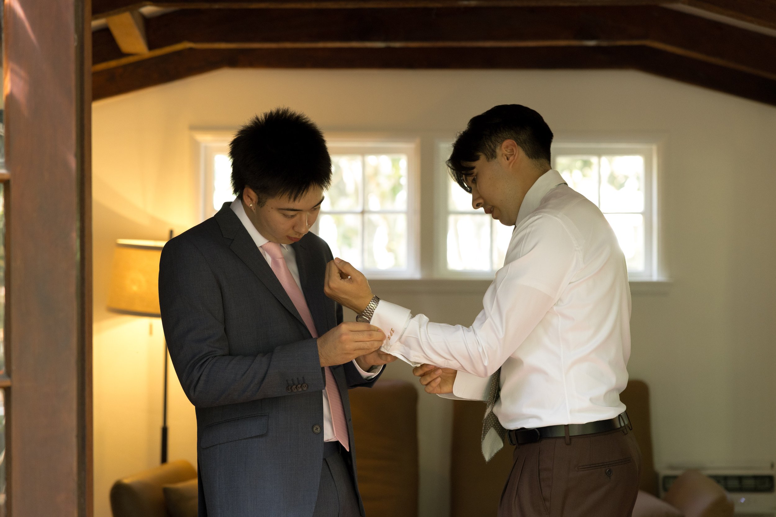 A groom and his groomsman, one in a dark suit and pink tie, and the other in a white shirt, are inside a room with wooden beams and windows, with one man adjusting the other's cufflink at a Rancho Las Lomas Wedding in Los Angeles