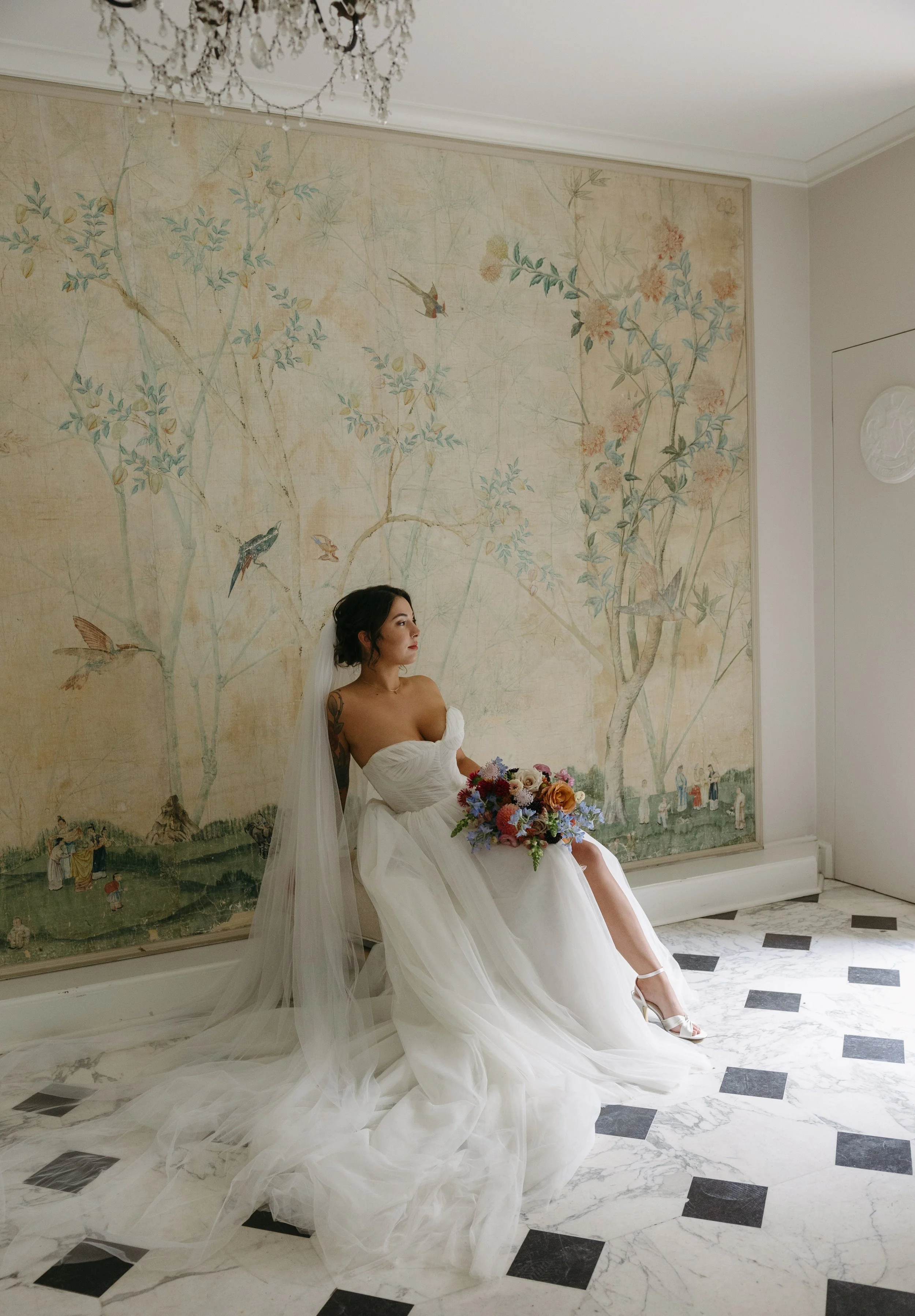 A bride sitting against a wall with a mural, holding a colorful bouquet, dressed in a white strapless wedding gown with a long veil, in a room with marble flooring and ornate chandelier.