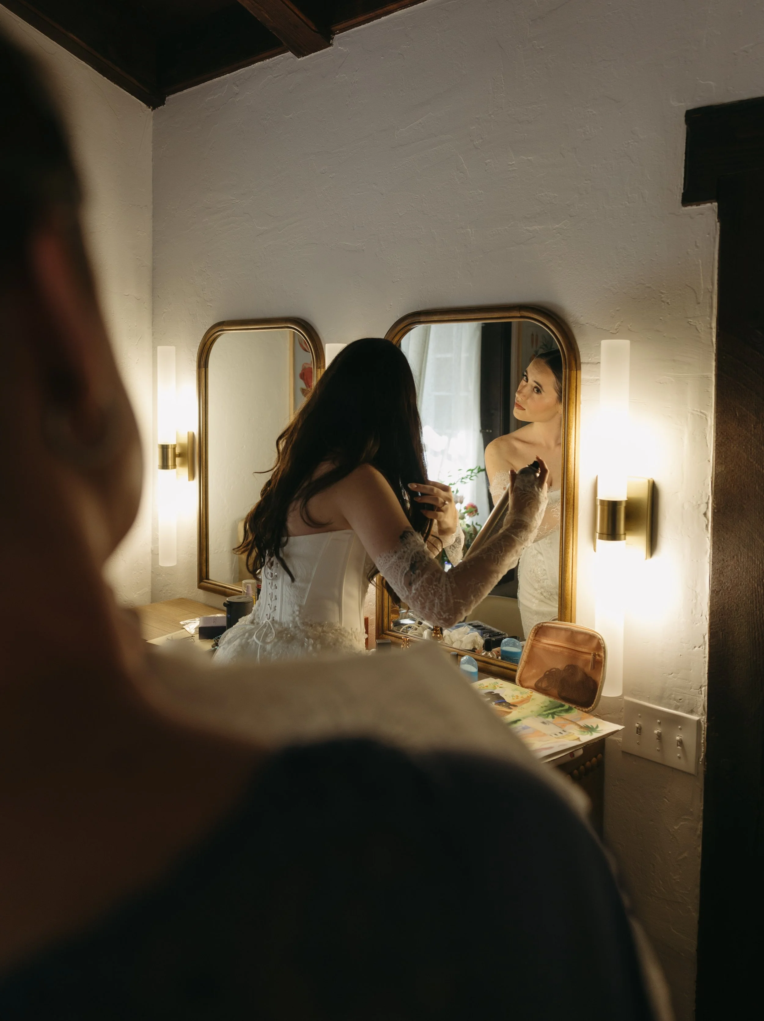 A woman in wedding attire looking at herself in a mirror in a warmly lit room, with her reflection seen in the mirror.