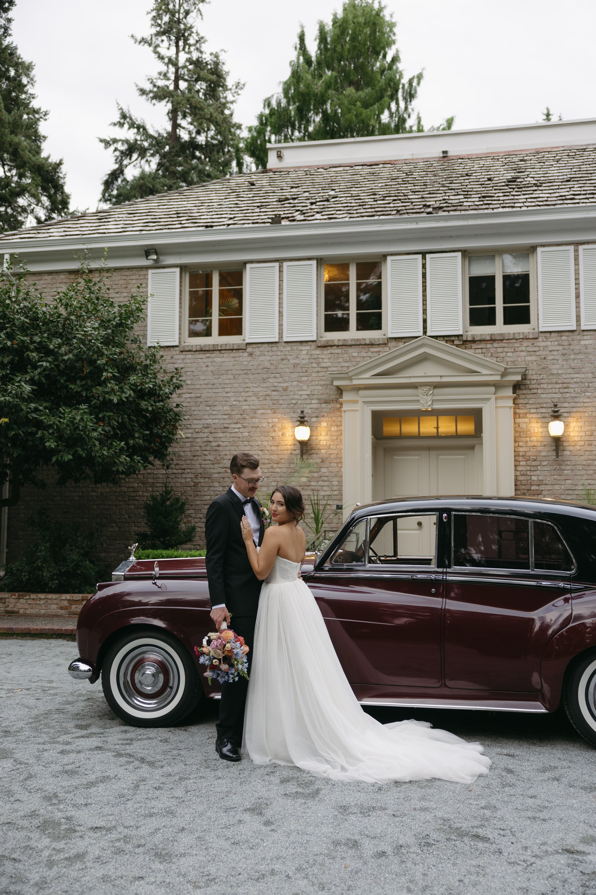 A bride and groom in wedding attire standing next to a vintage car in front of a brick house with white shutters at a European inspired Lakewold Gardens wedding in Seattle