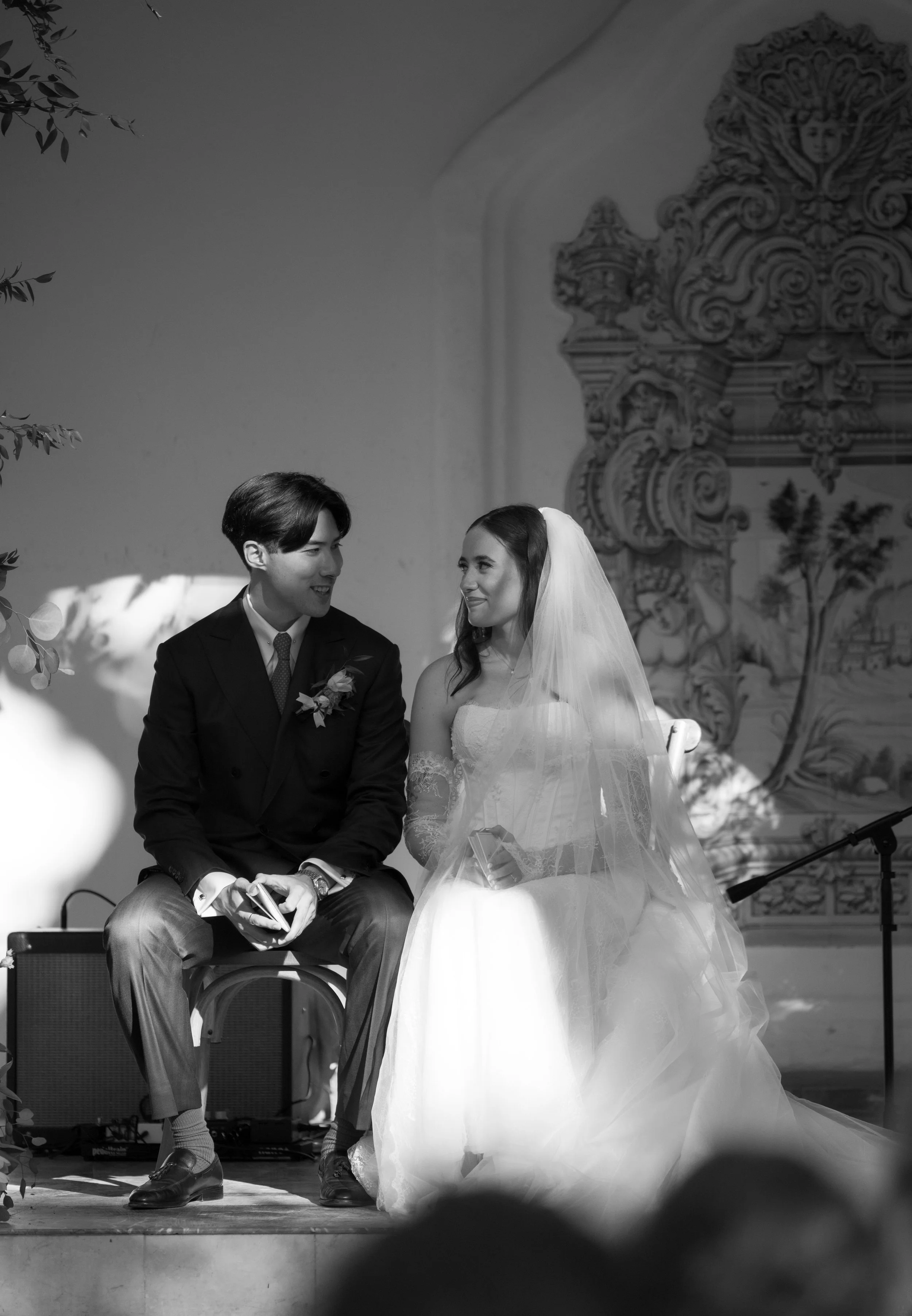 A black-and-white photo of a wedding ceremony, featuring a bride and groom sitting on stage. The bride is wearing a wedding gown with a veil and the groom is dressed in a suit. They are looking at each other and smiling.