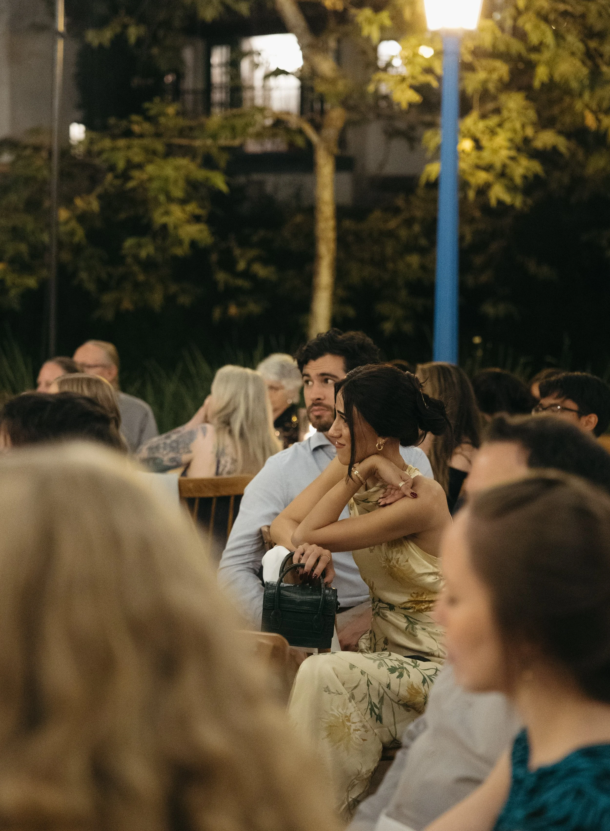 A group of people seated outdoors at night, some engaging in conversation, with trees and a blue lamppost in the background.