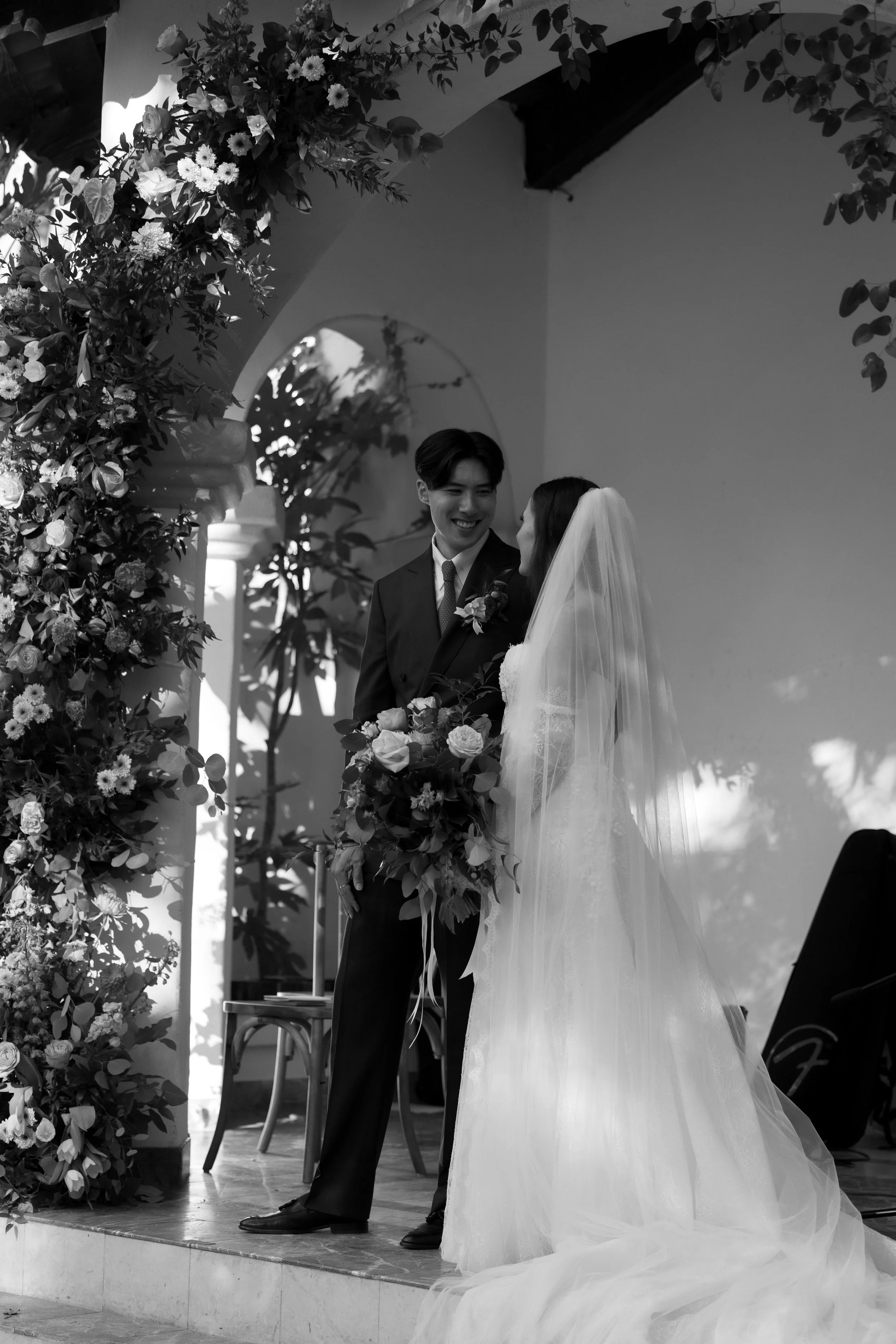 A bride and groom smiling at each other during their wedding ceremony, standing under an arch decorated with flowers, with a church-like setting in the background.