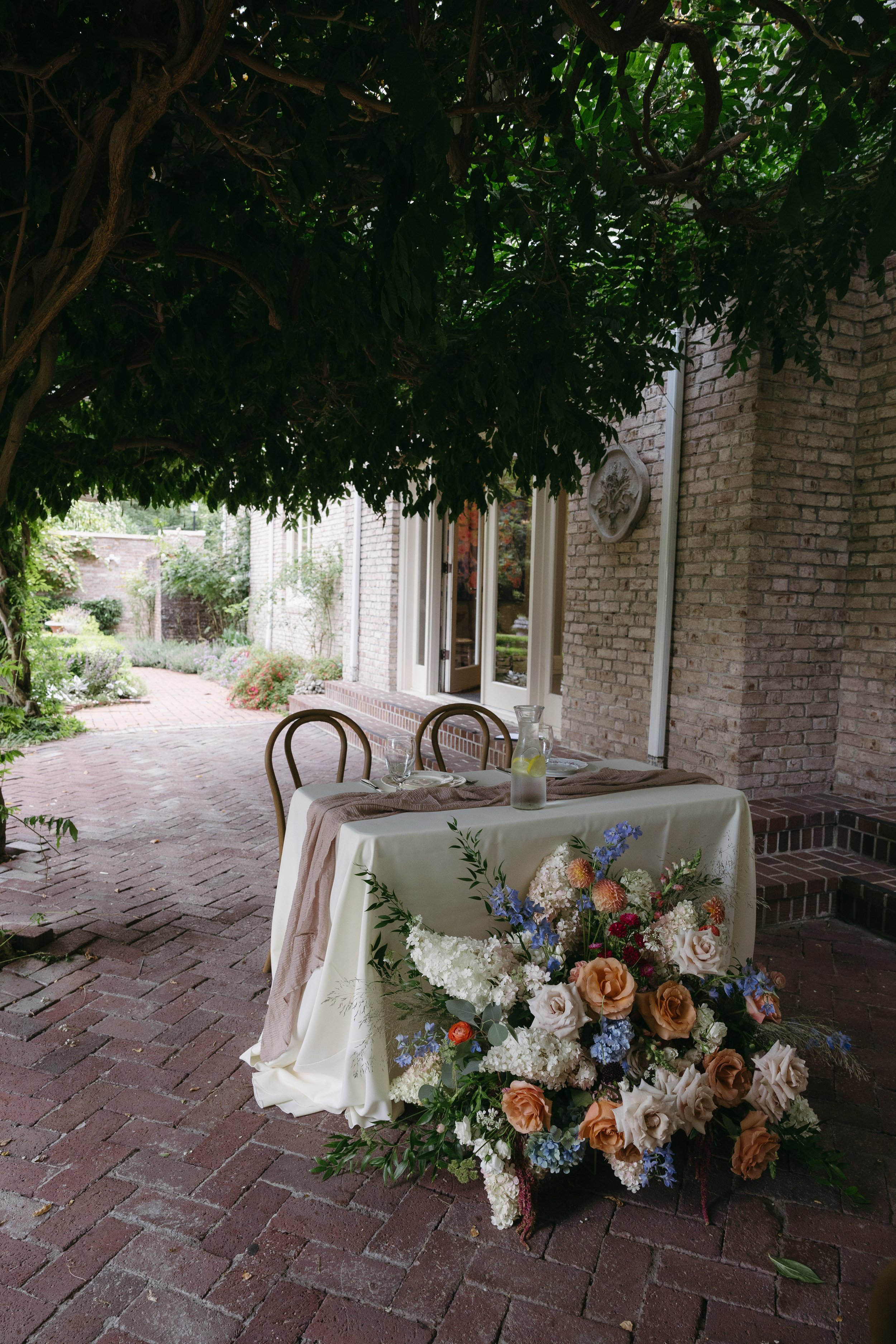 A decorated outdoor table with a floral arrangement, set for dining under a tree, on a brick patio adjacent to a brick house.