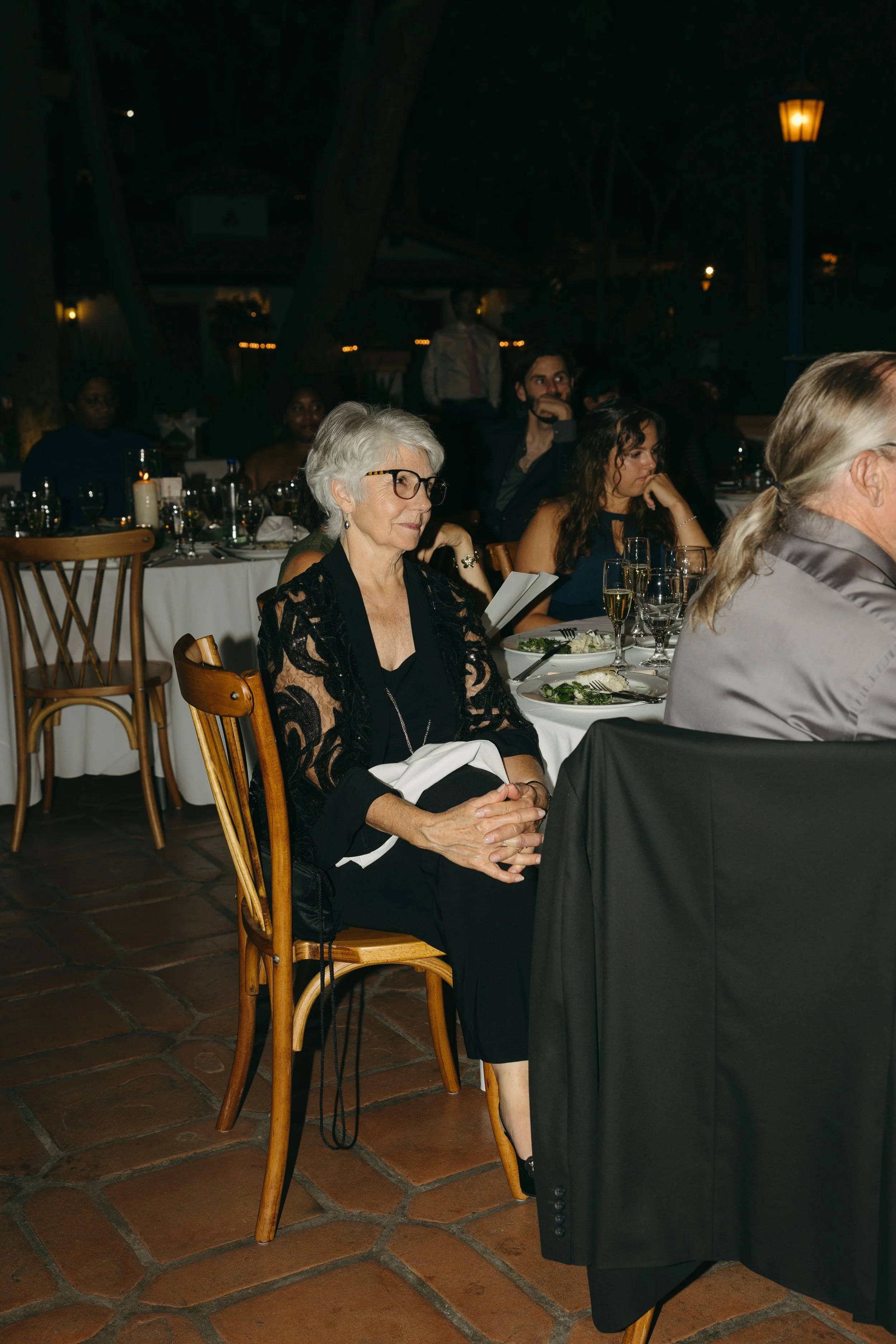 An older woman with gray hair and glasses is sitting at a formal dinner event, wearing a black outfit with a lace shawl, surrounded by other elegantly dressed guests at round tables with fine dining settings.