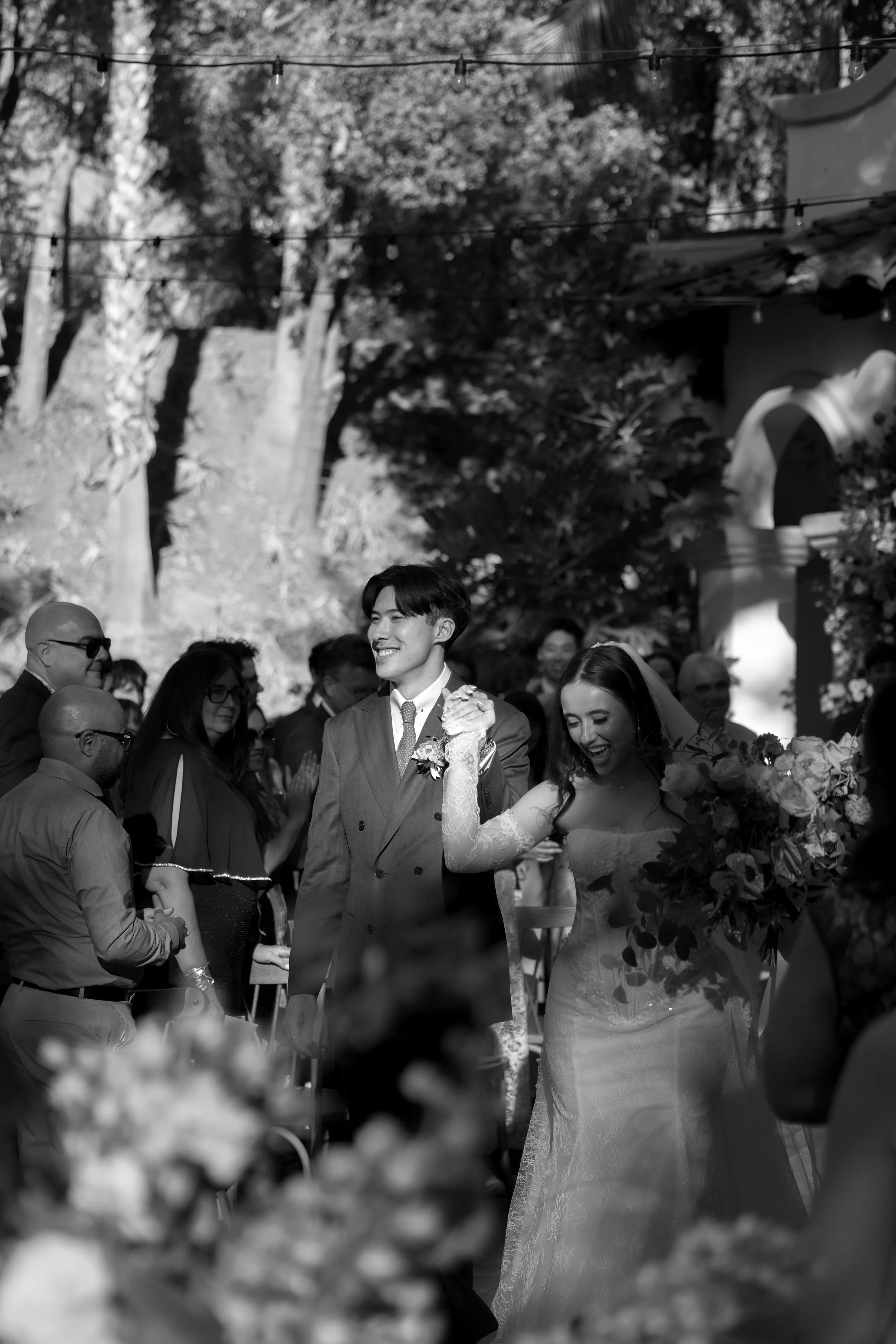 A bride and groom smiling and holding hands at an outdoor wedding ceremony, surrounded by guests at a Rancho Las Lomas Wedding in Los Angeles