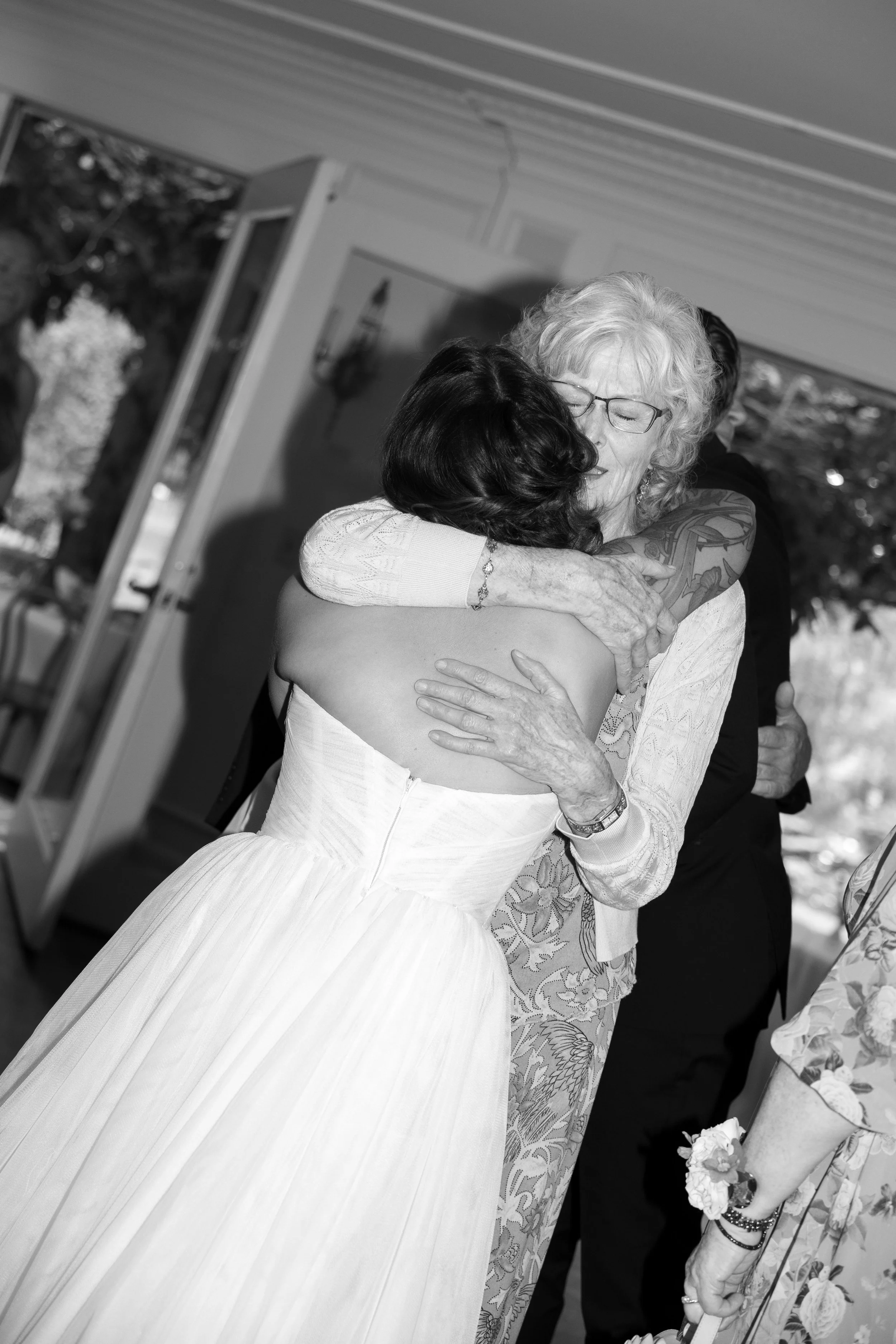 A bride hugging an elderly woman, possibly her grandmother, at a wedding reception in a room with large windows and trees outside.
