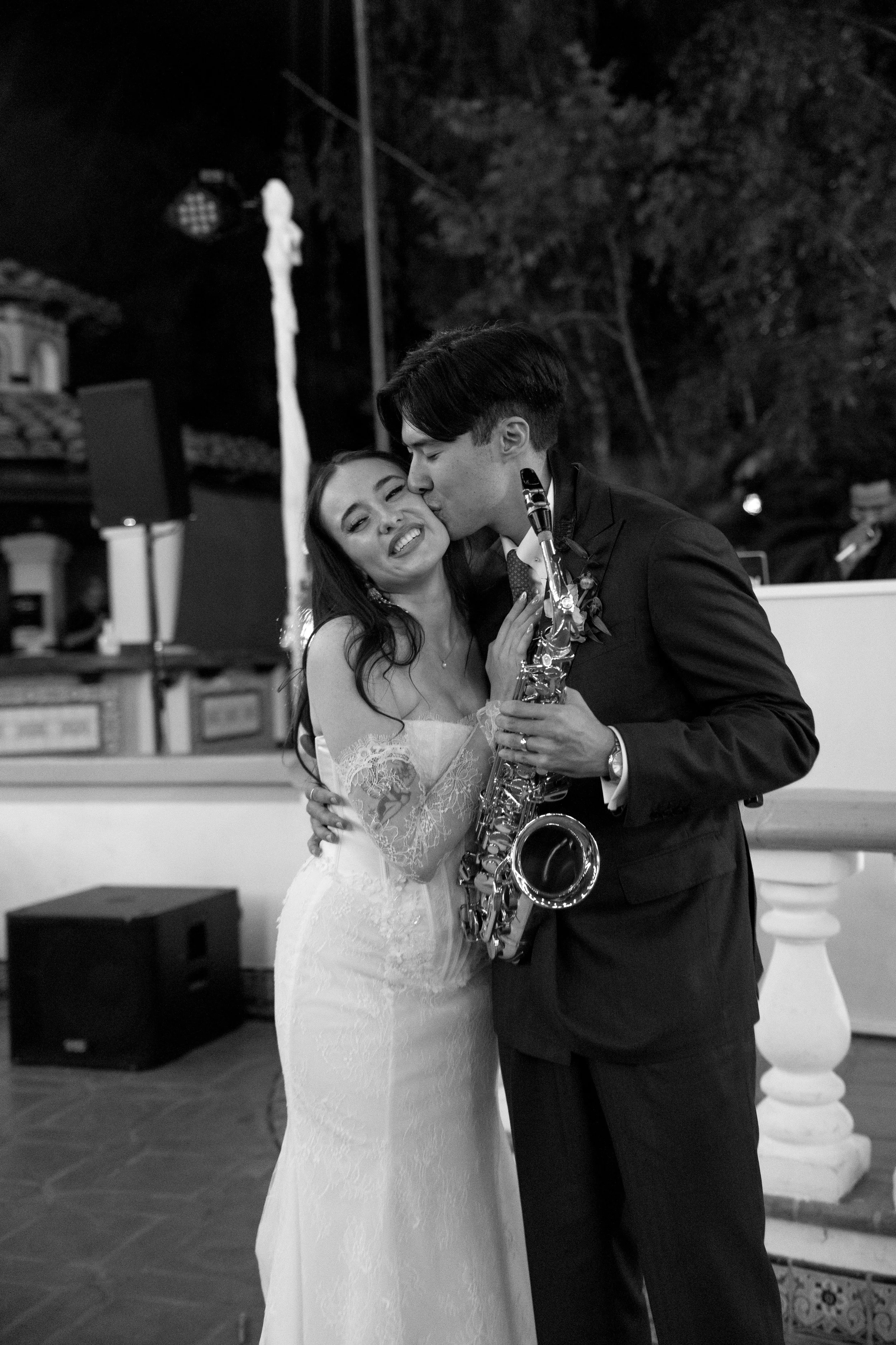 A black-and-white photo of a newlywed couple at their wedding, with the groom playing a saxophone. The groom is kissing the bride on the cheek while she smiles with her eyes closed, embracing him.