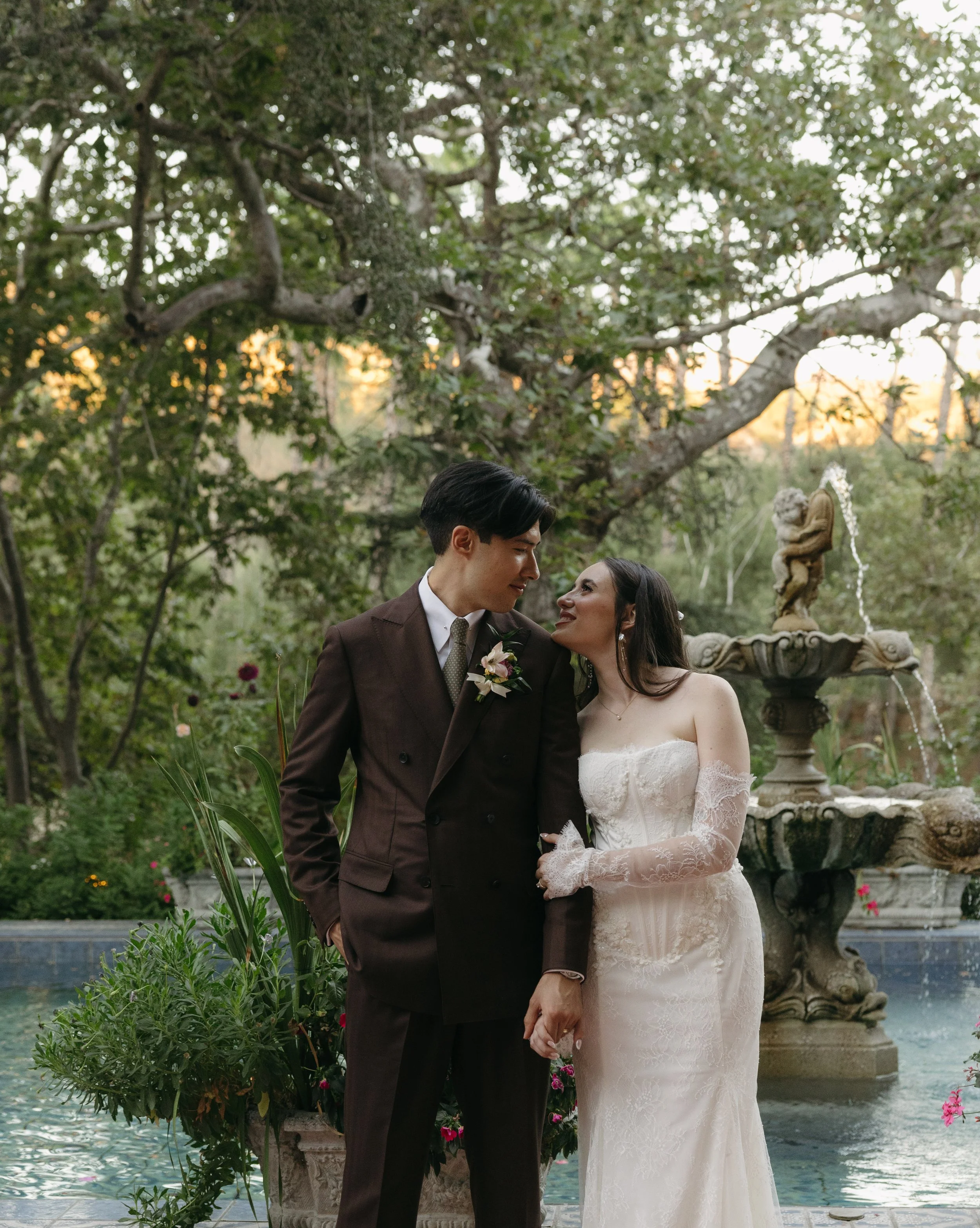 A couple in wedding attire stands near a fountain in a garden at sunset, gazing at each other affectionately at a Rancho Las Lomas Wedding in Los Angeles