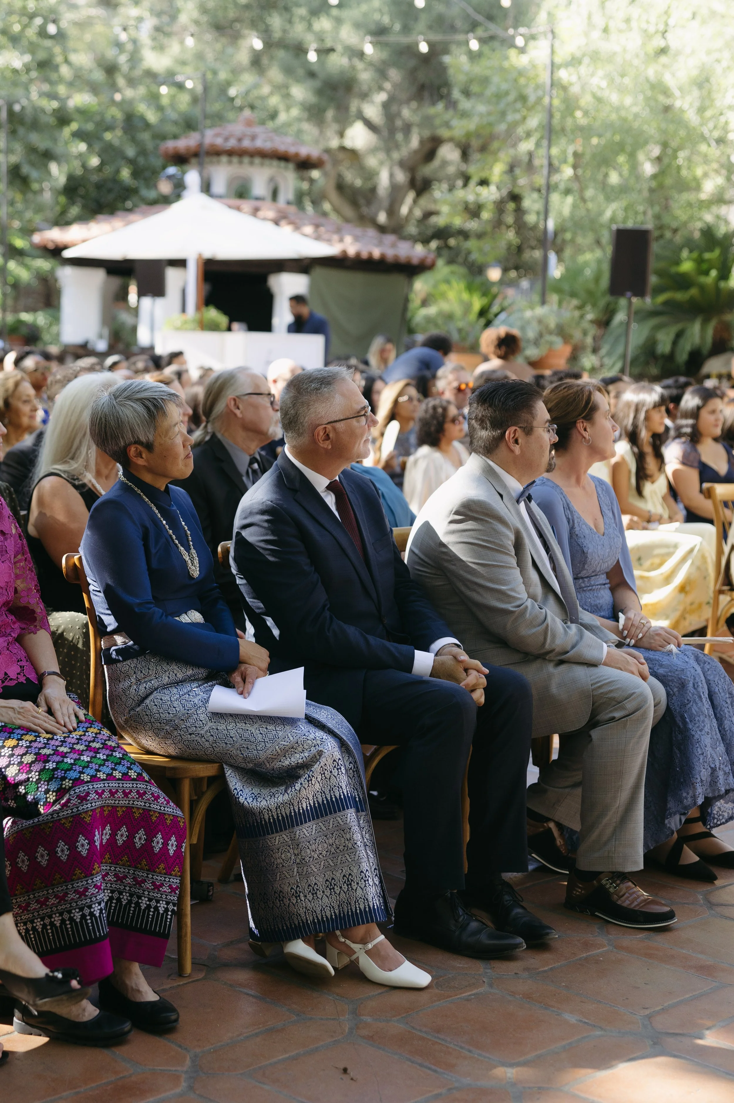 People seated outdoors at a formal event or ceremony, with a decorative building and greenery in the background at a Rancho Las Lomas Wedding in Los Angeles