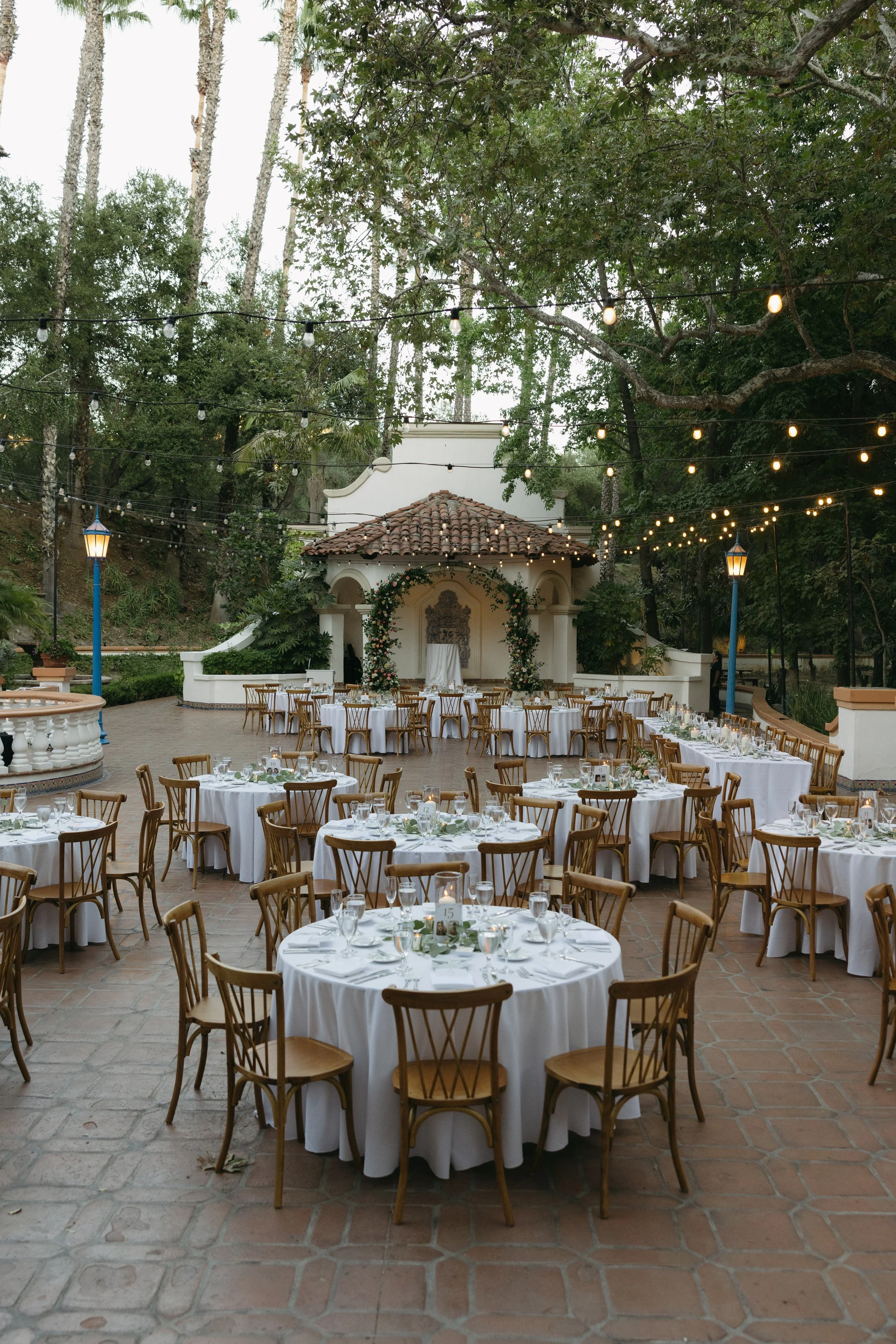 Outdoor event space set up with round tables with white tablecloths, wooden chairs, and table settings, decorated with candles and greenery, surrounded by trees and string lights at a Rancho Las Lomas Wedding in Los Angeles