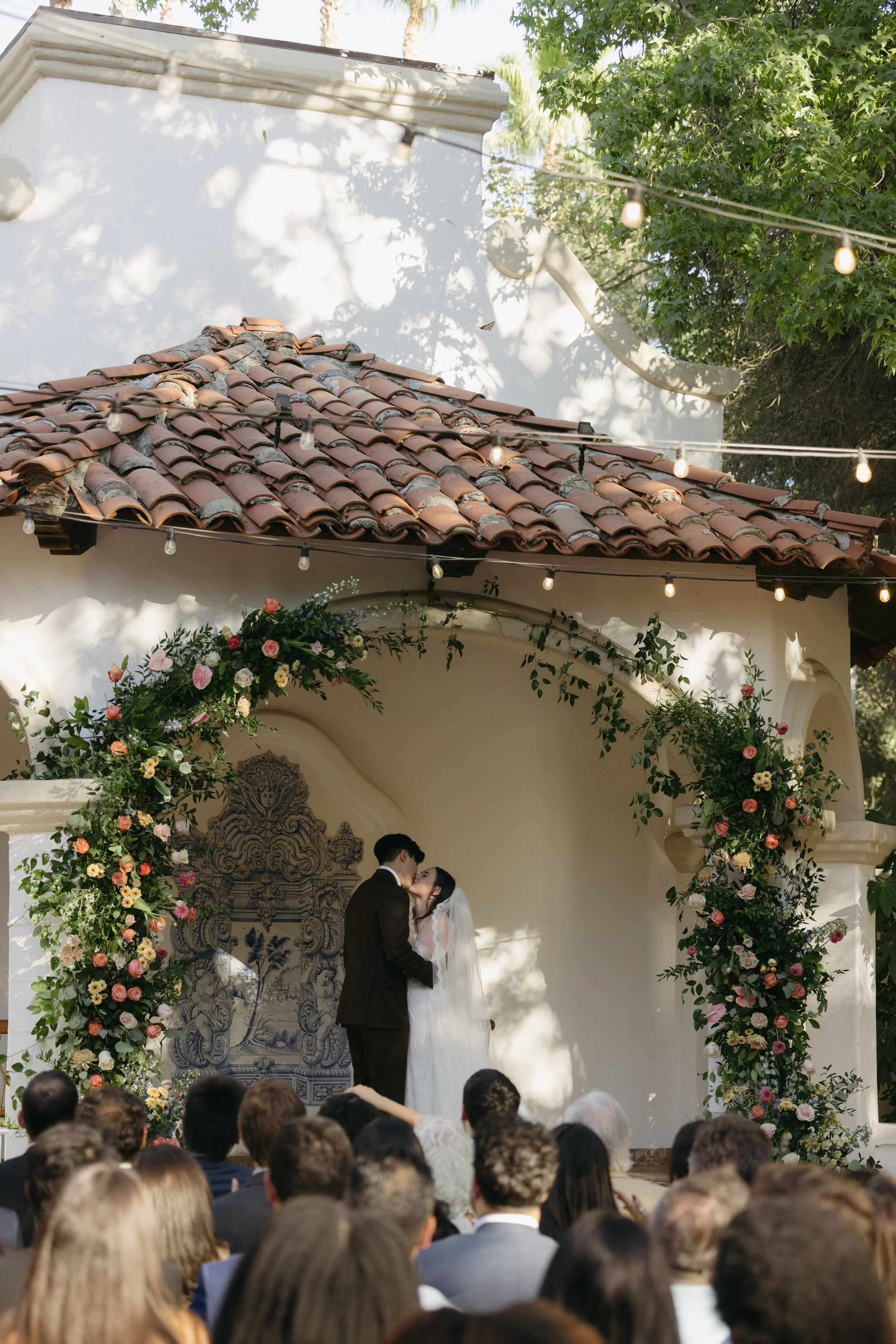 A bride and groom kissing during their outdoor wedding ceremony under a floral arch with an audience watching at a Rancho Las Lomas Wedding in Los Angeles
