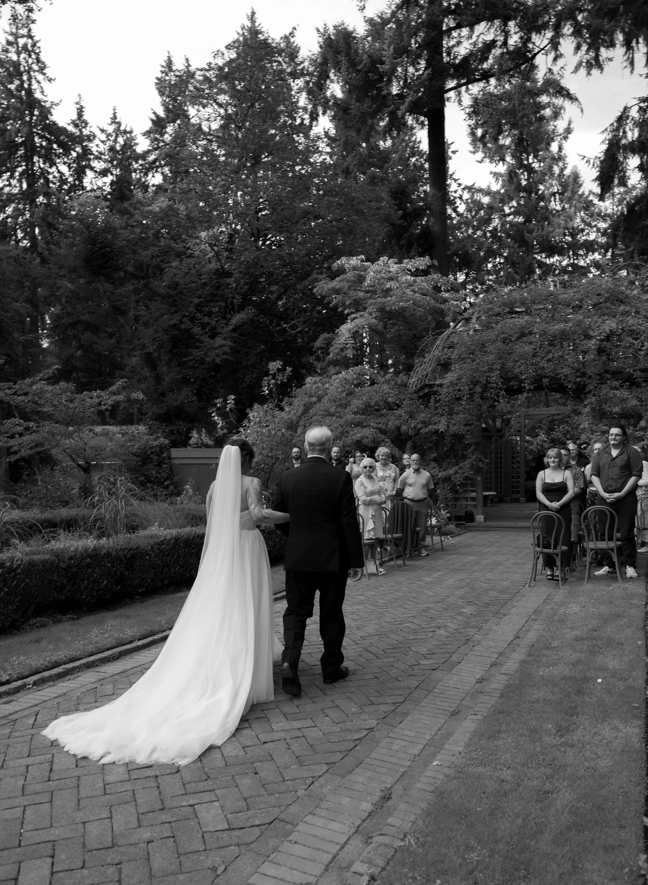 A black and white photo of a wedding ceremony outdoors, with a bride in a long train and a man, likely her father, walking her down a garden path, facing a group of seated and standing guests.