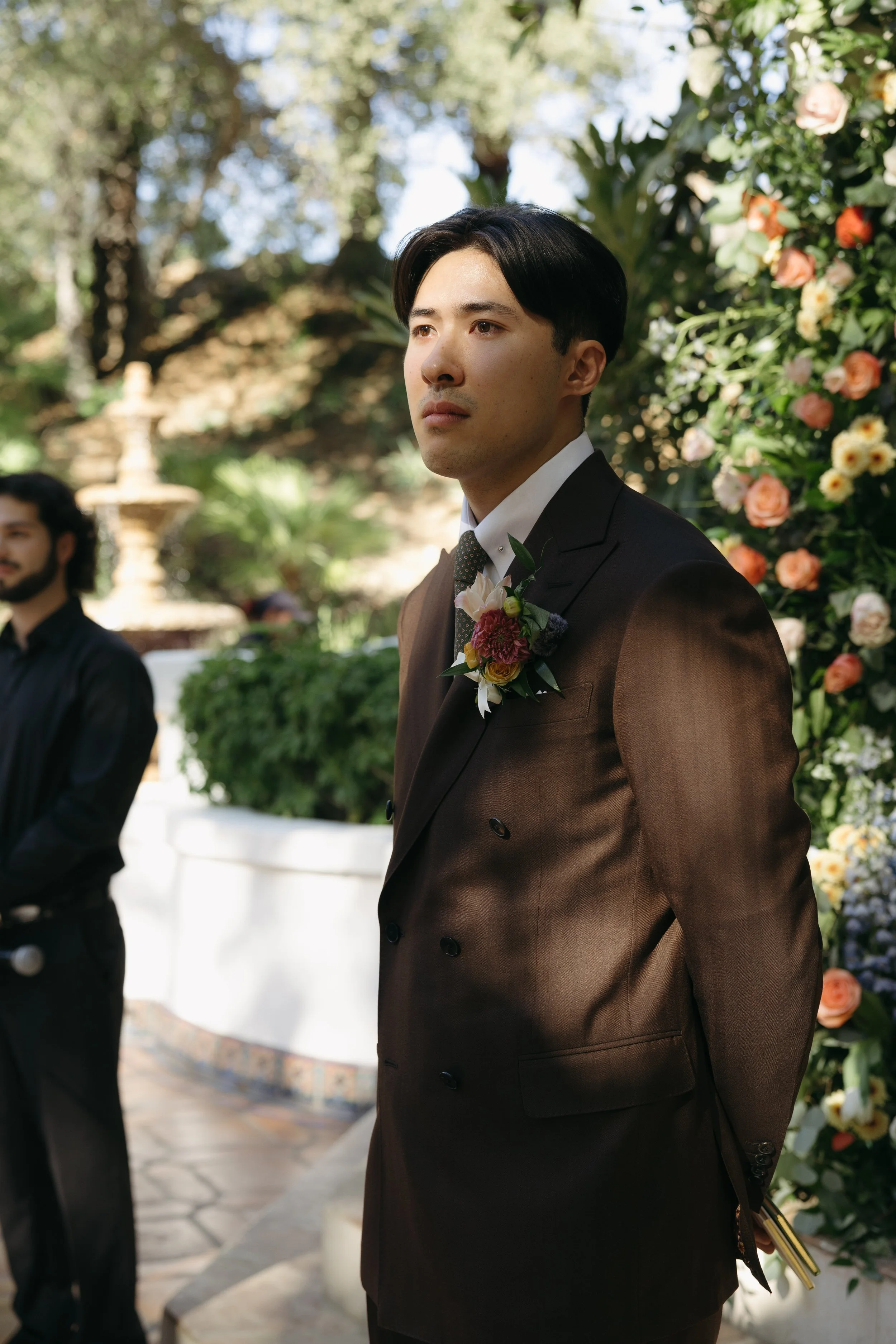 A man in a dark brown suit with a floral boutonniere on his lapel, standing outdoors during a formal event, possibly a wedding, with a floral backdrop and greenery.