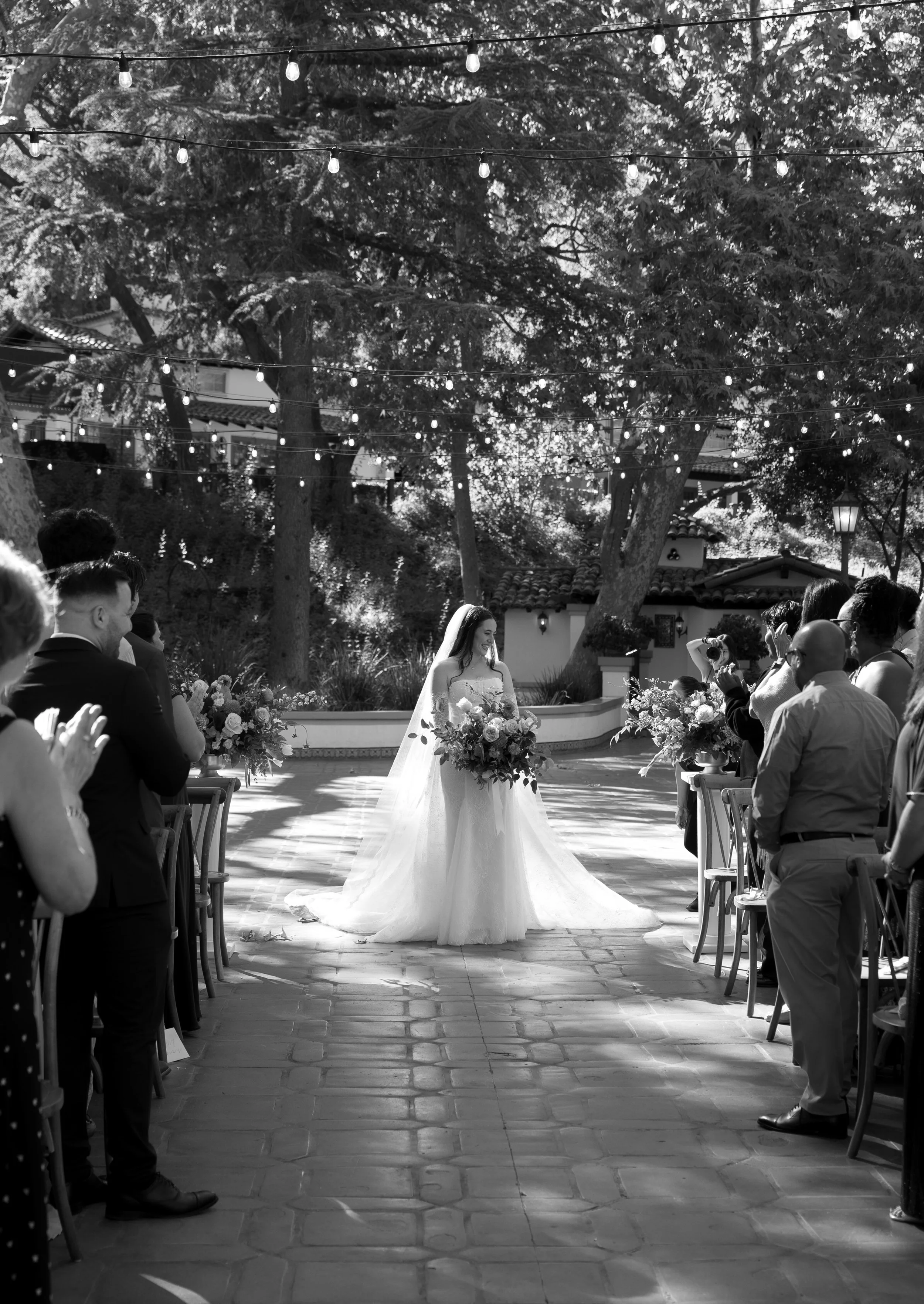 A bride in a wedding gown holding a bouquet stands under string lights at an outdoor wedding ceremony with guests on either side at a Rancho Las Lomas Wedding in Los Angeles