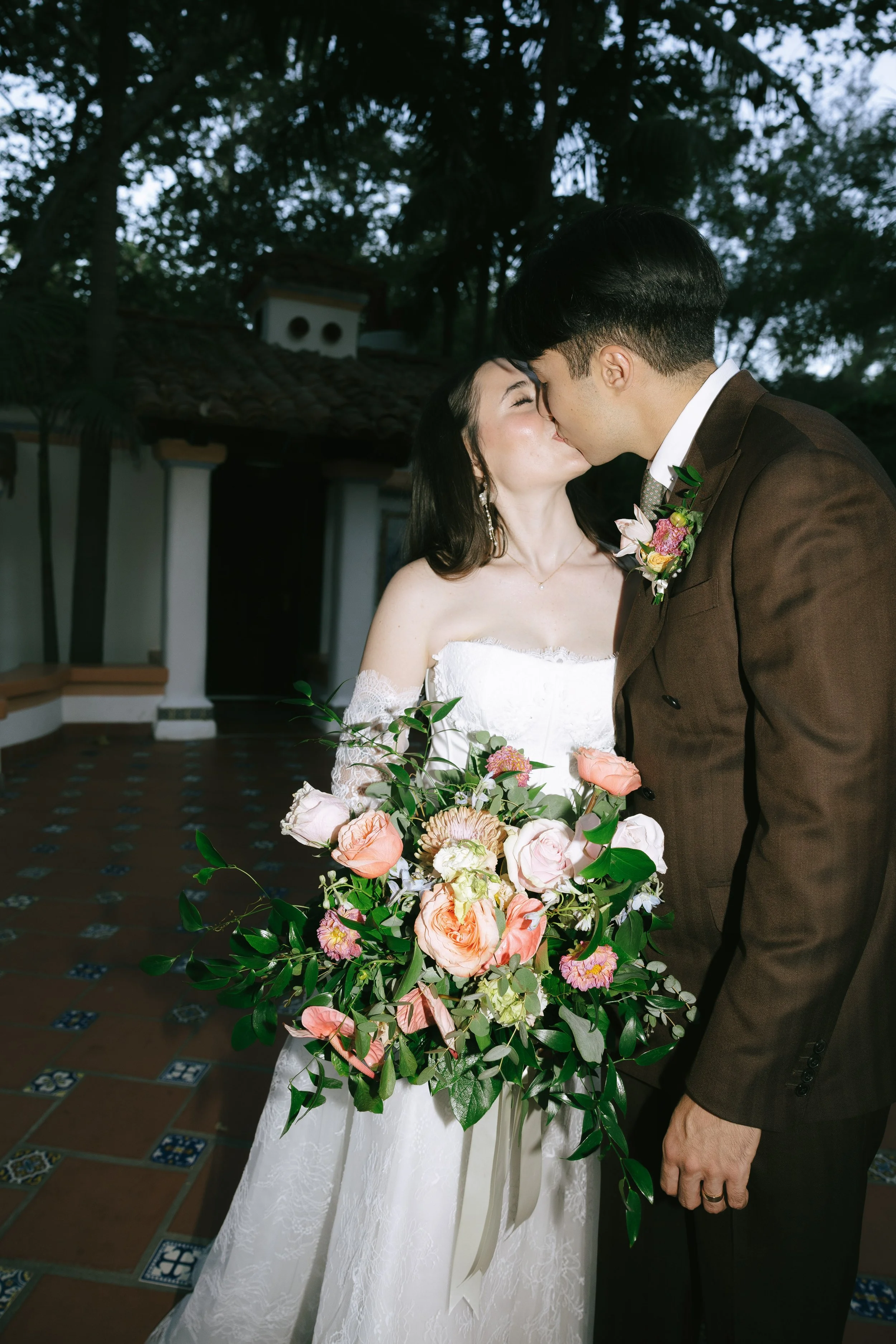 A bride and groom sharing a kiss outdoors, with the bride holding a large bouquet of pink, peach, and cream flowers, and a stone-tiled patio and greenery in the background.