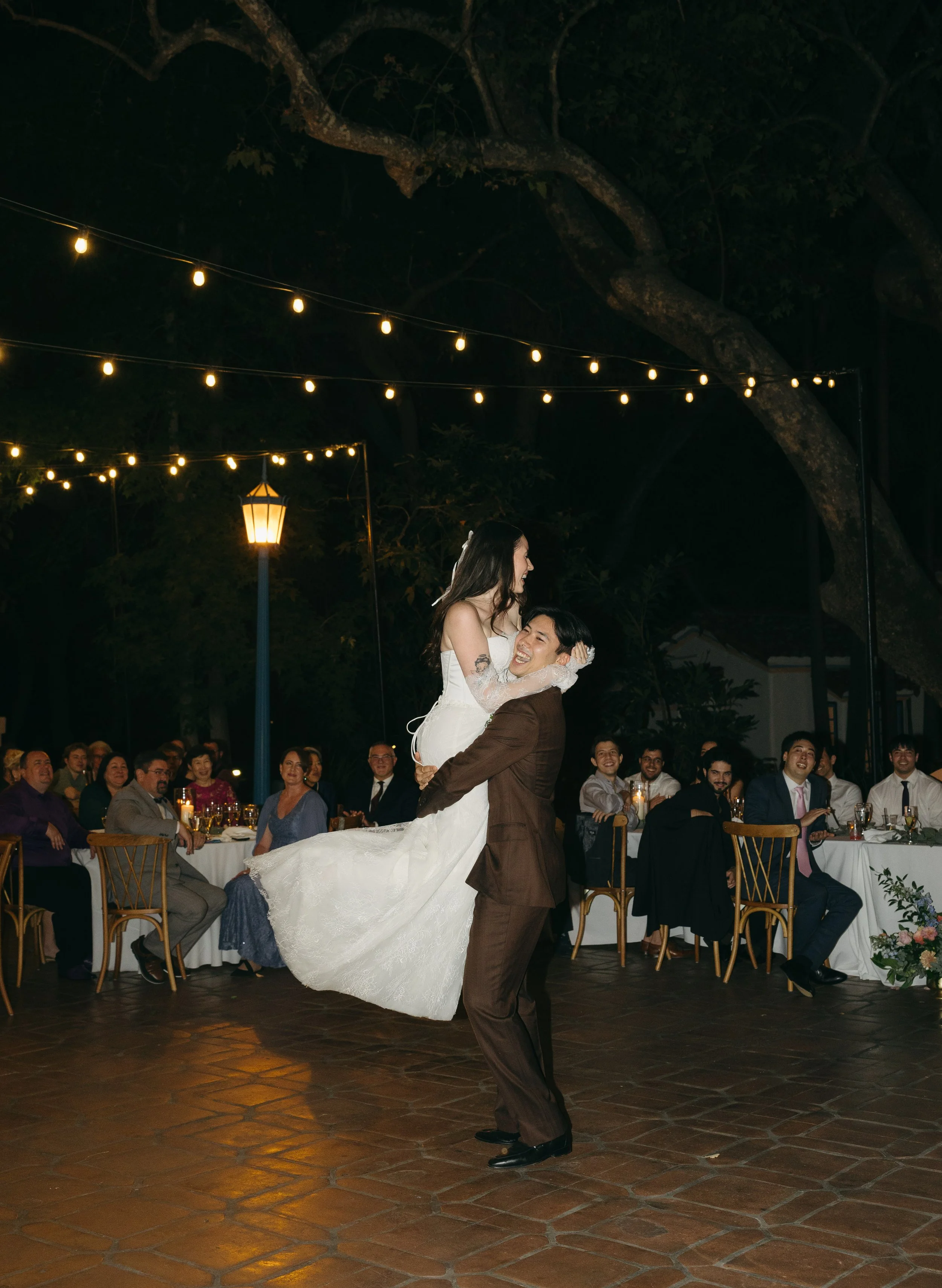 Man in a brown suit lifting a woman in a white wedding dress at night, outdoors, under string lights with seated guests watching, some smiling.