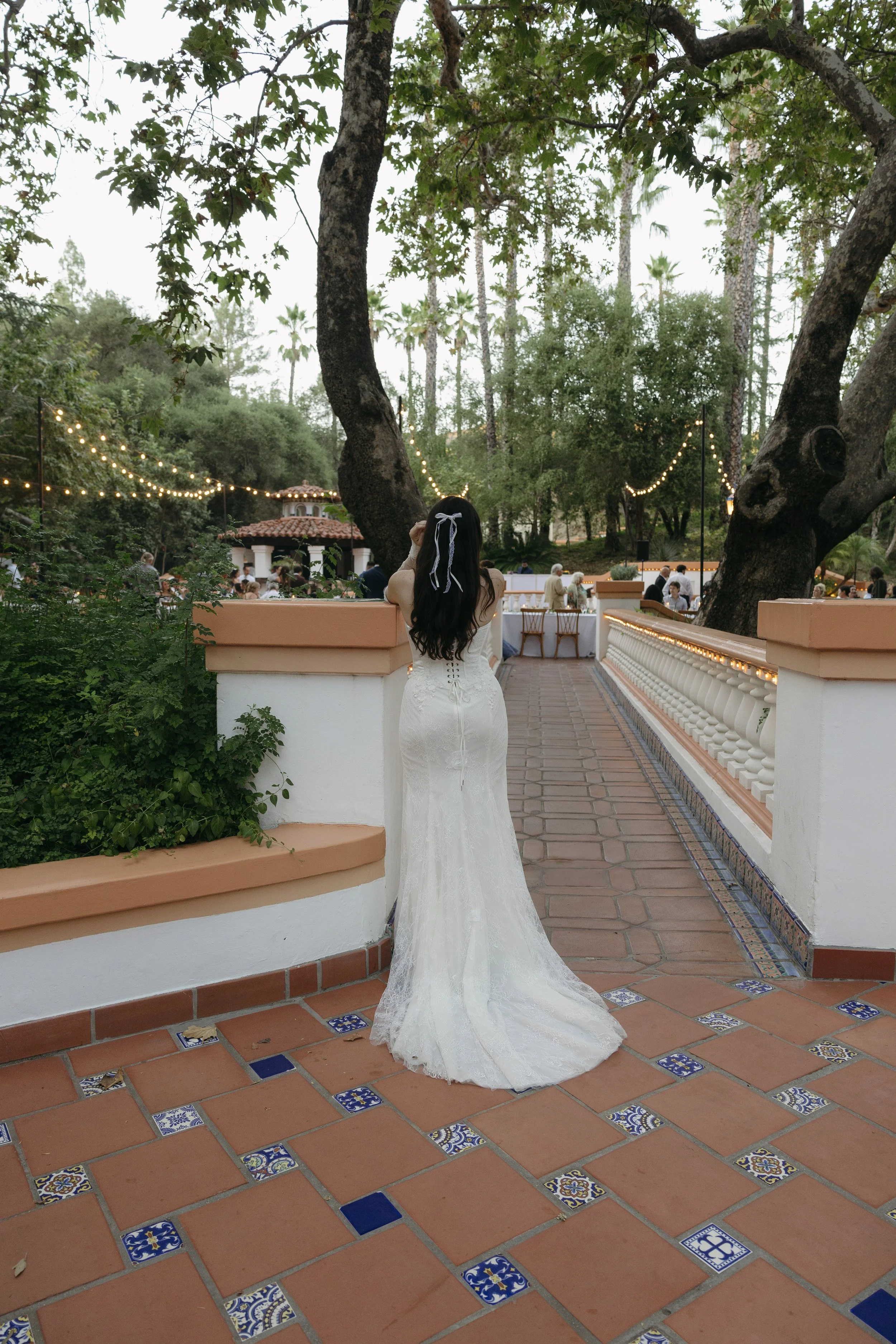 A woman in a white lace wedding dress with a ribbon in her dark, long hair stands on a terracotta tiled patio during an outdoor event, facing away with a garden and string lights in the background.