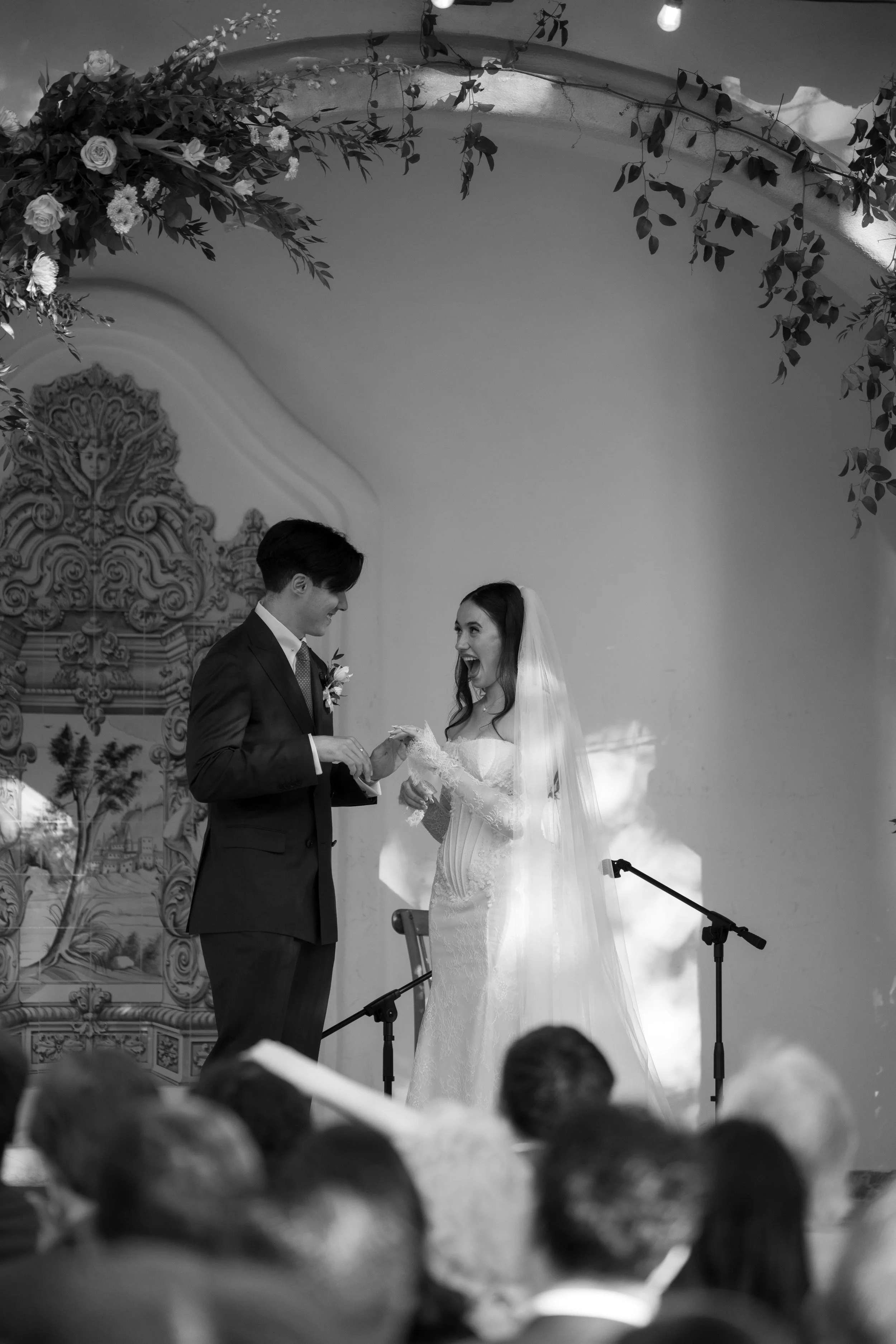 Black and white photo of a bride and groom exchanging rings during their wedding ceremony, with guests watching in the foreground and decorative floral and ornate backdrop behind them.