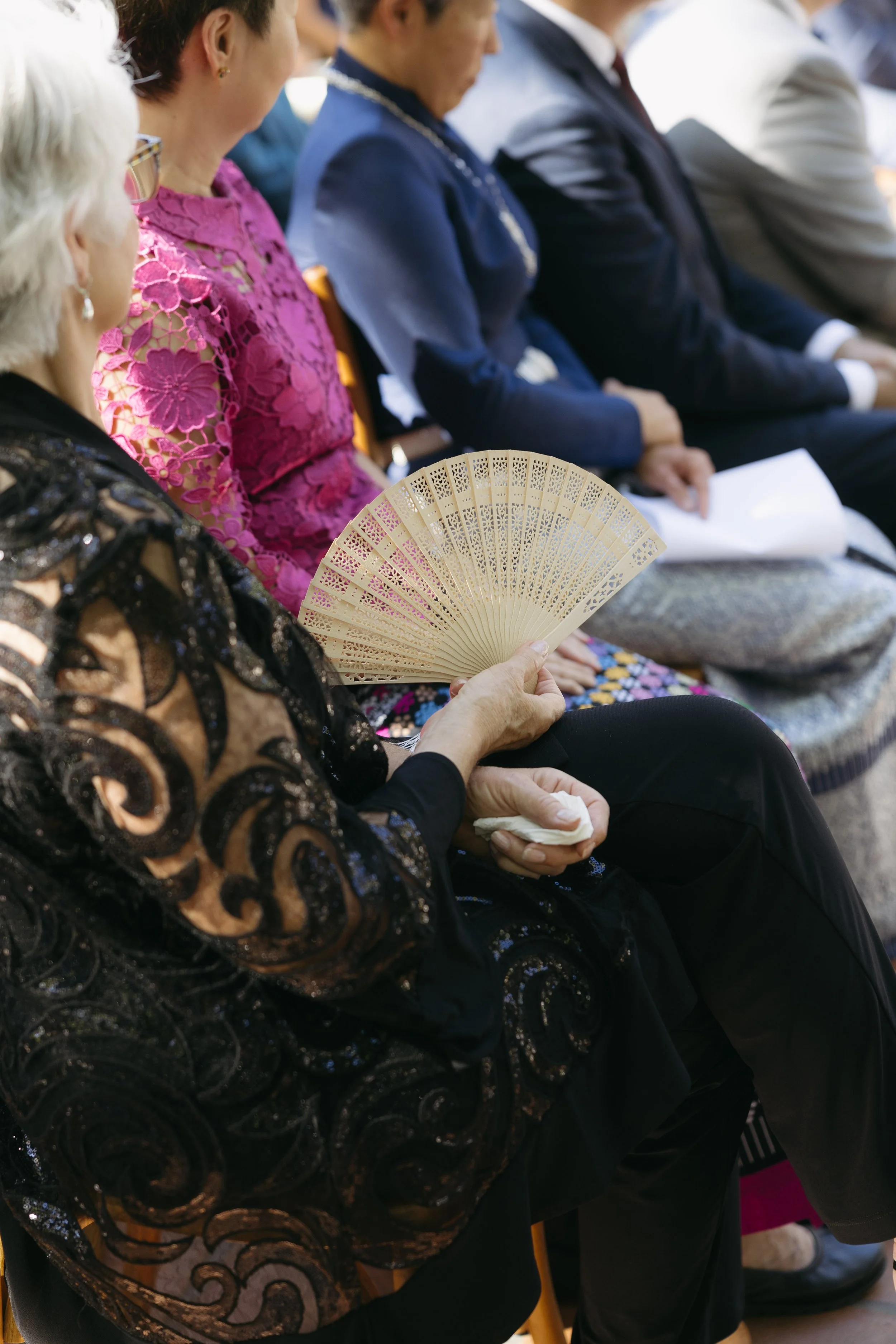 A group of people sitting in a row, dressed in formal and colorful attire, with one person holding an ornate fan.