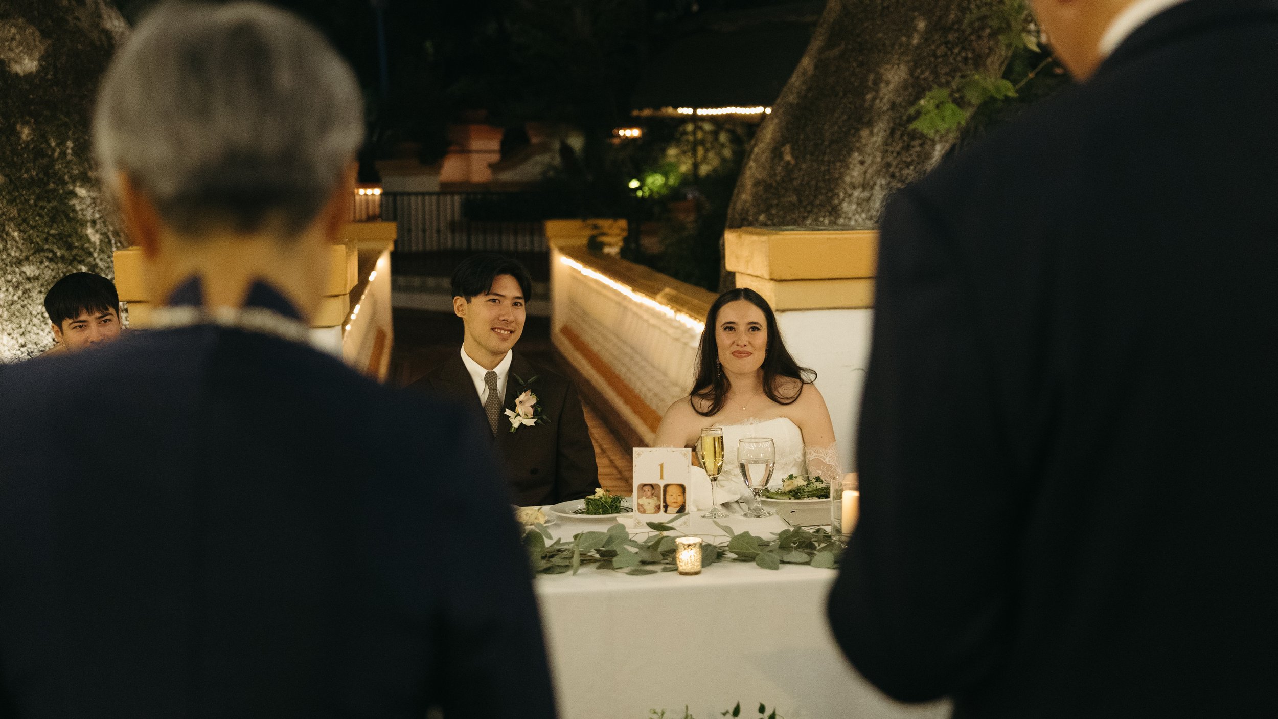 A bride and groom sitting at a wedding reception table, listening to speeches, with guests in the foreground. It is evening with outdoor lighting.