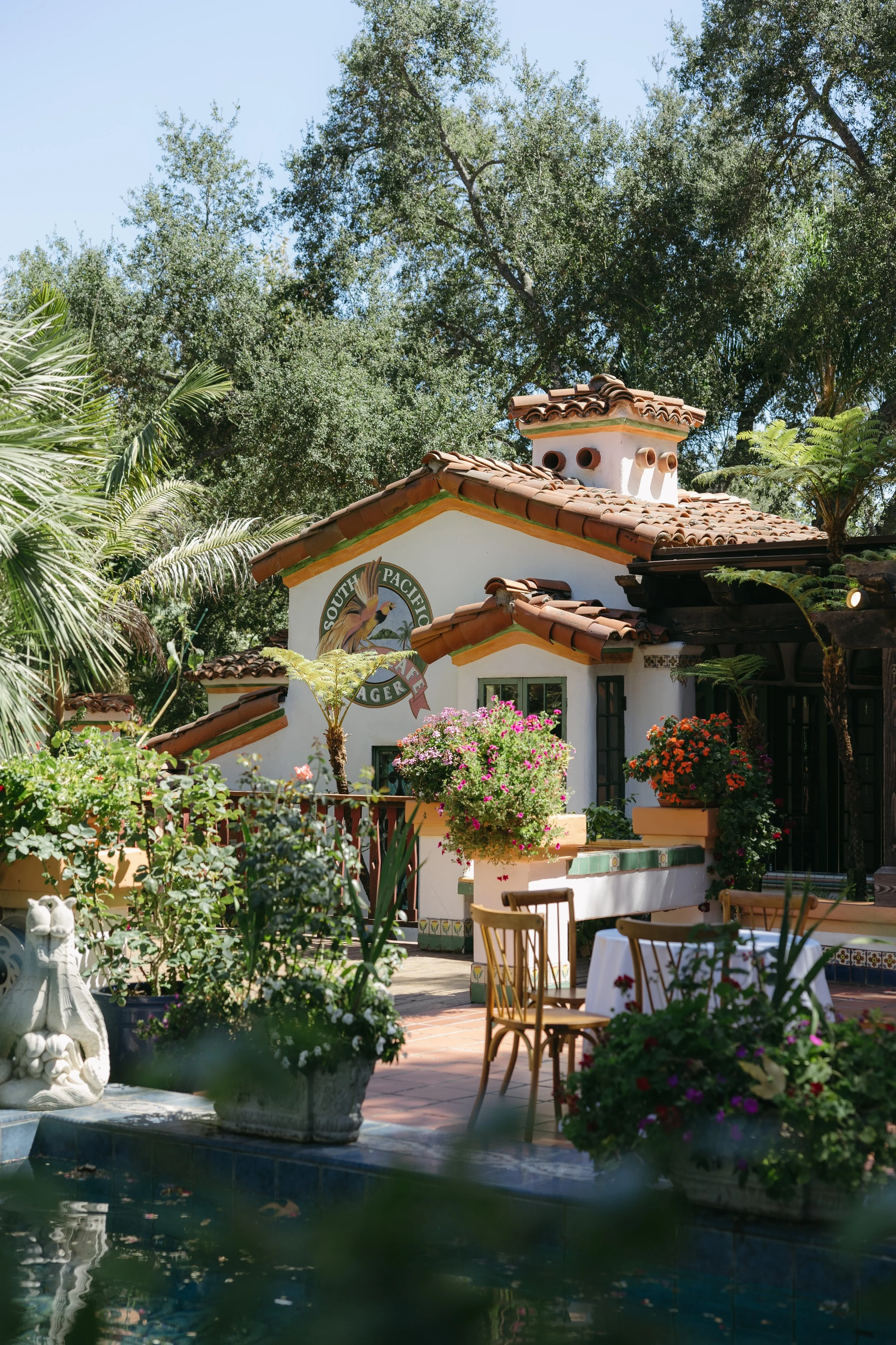 A charming Spanish-style building with a terracotta-tiled roof, surrounded by lush greenery and colorful potted flowers, including pink and orange blooms, with outdoor seating and a pond in the foreground at a Rancho Las Lomas Wedding in Los Angeles