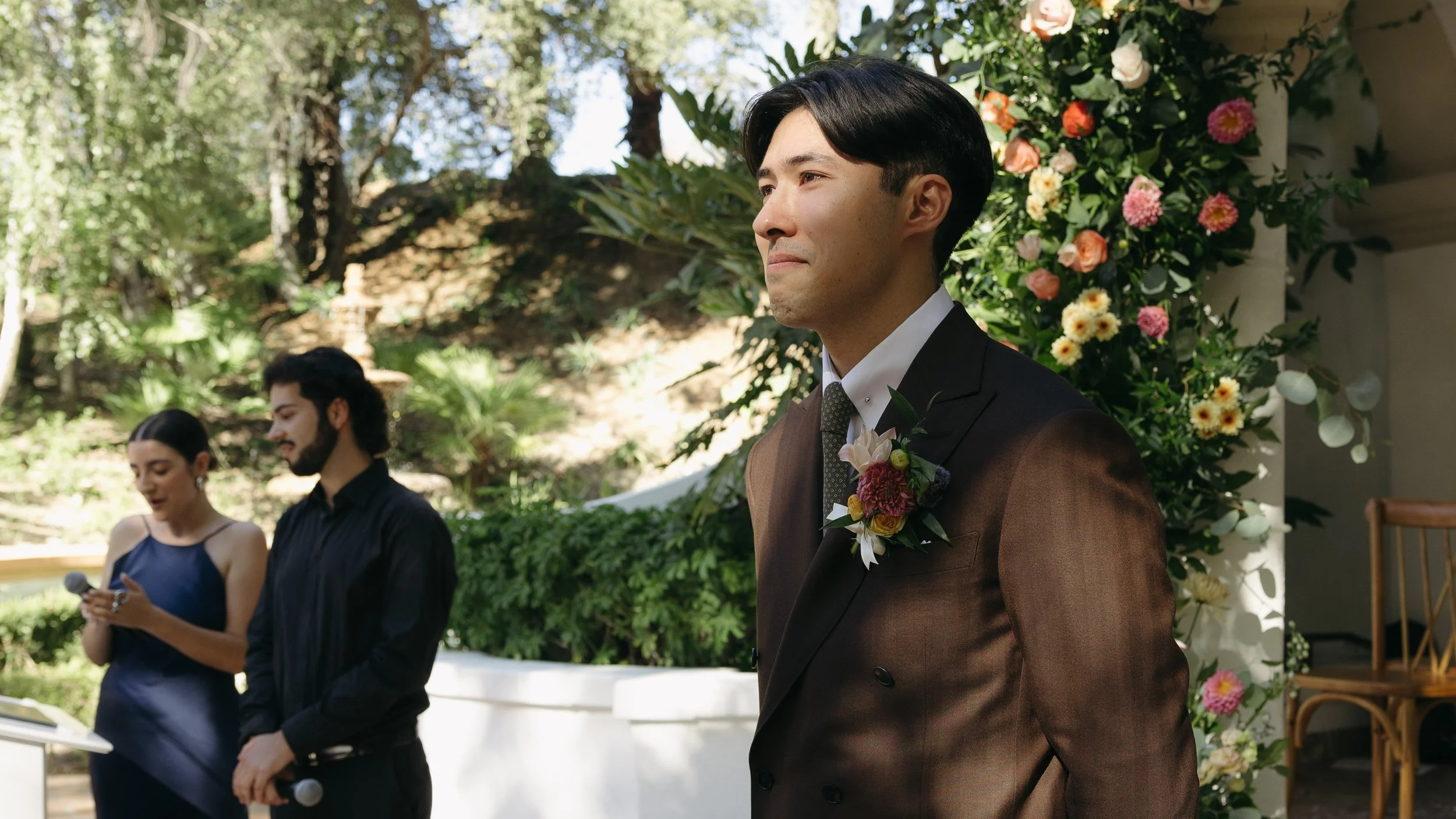 A man in a brown suit with a boutonnière stands outdoors at a wedding ceremony, next to a floral backdrop. In the background, a woman in a navy dress and a man in a black shirt hold a microphone and a phone, respectively, under dappled sunlight.
