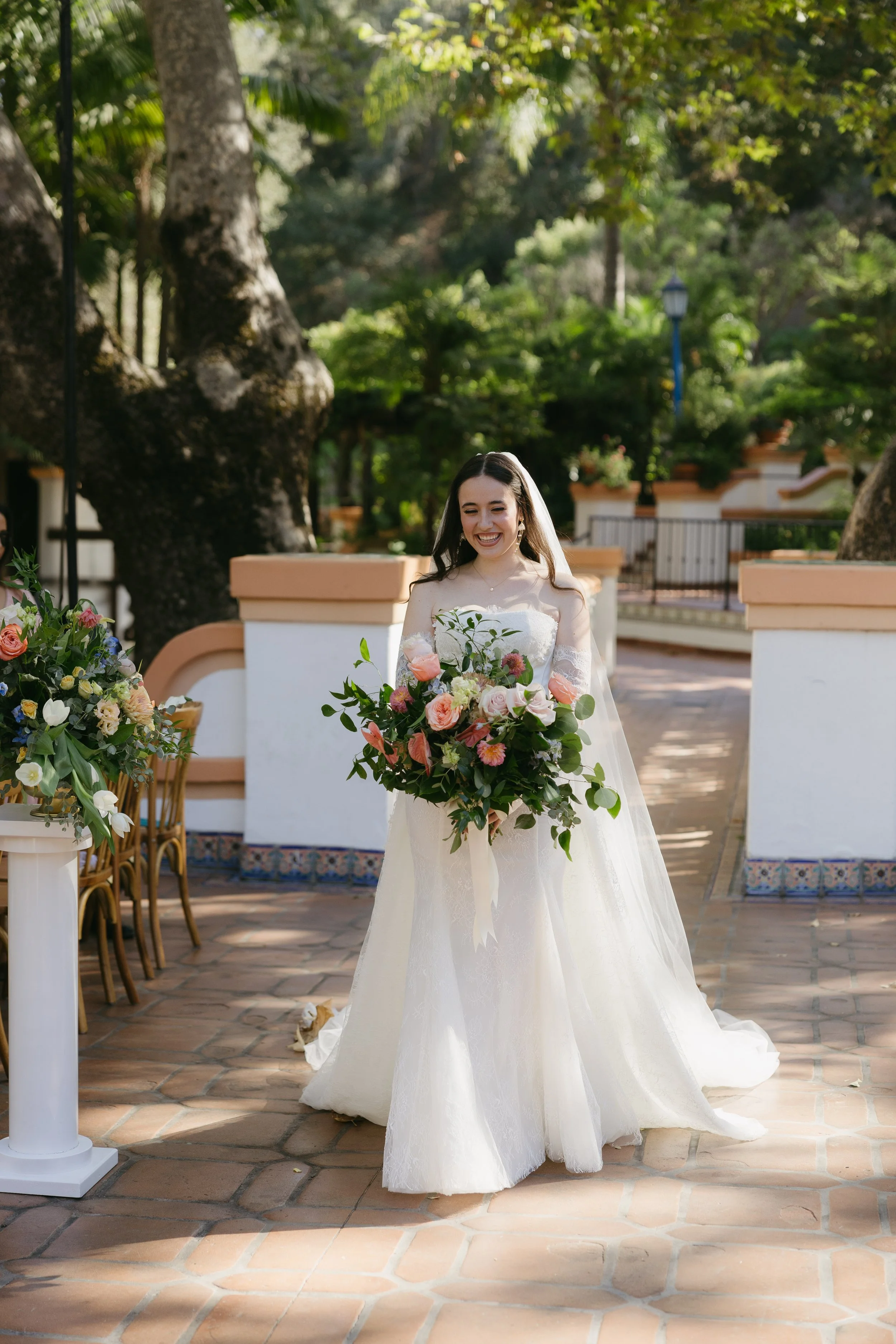 A bride in a white wedding dress holding a large bouquet of pink, white, and green flowers, standing outdoors on a brick patio surrounded by trees and greenery, smiling.