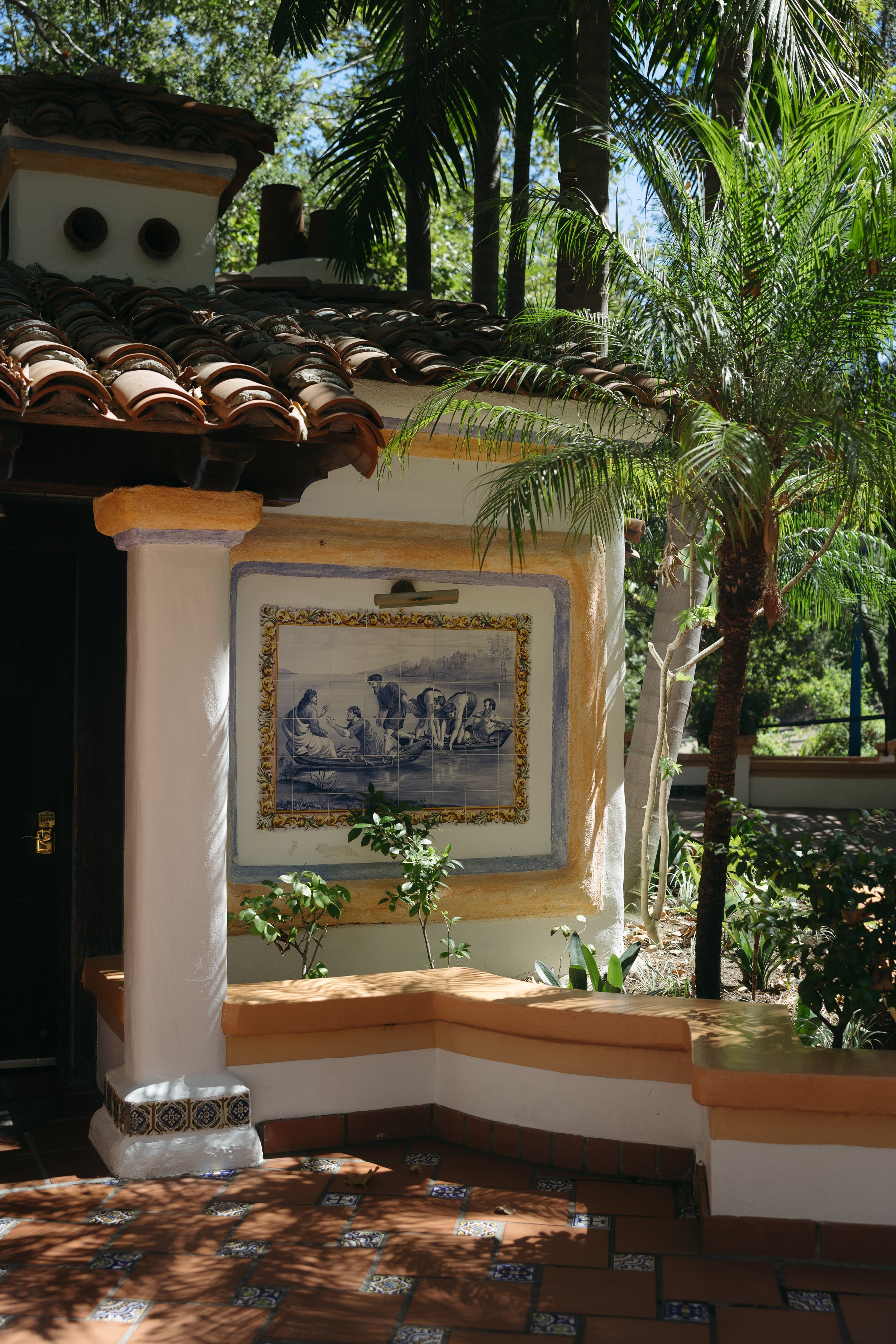 A tiled patio with decorative tiles, a stucco wall with a framed tile artwork, and lush green plants and trees providing shade.