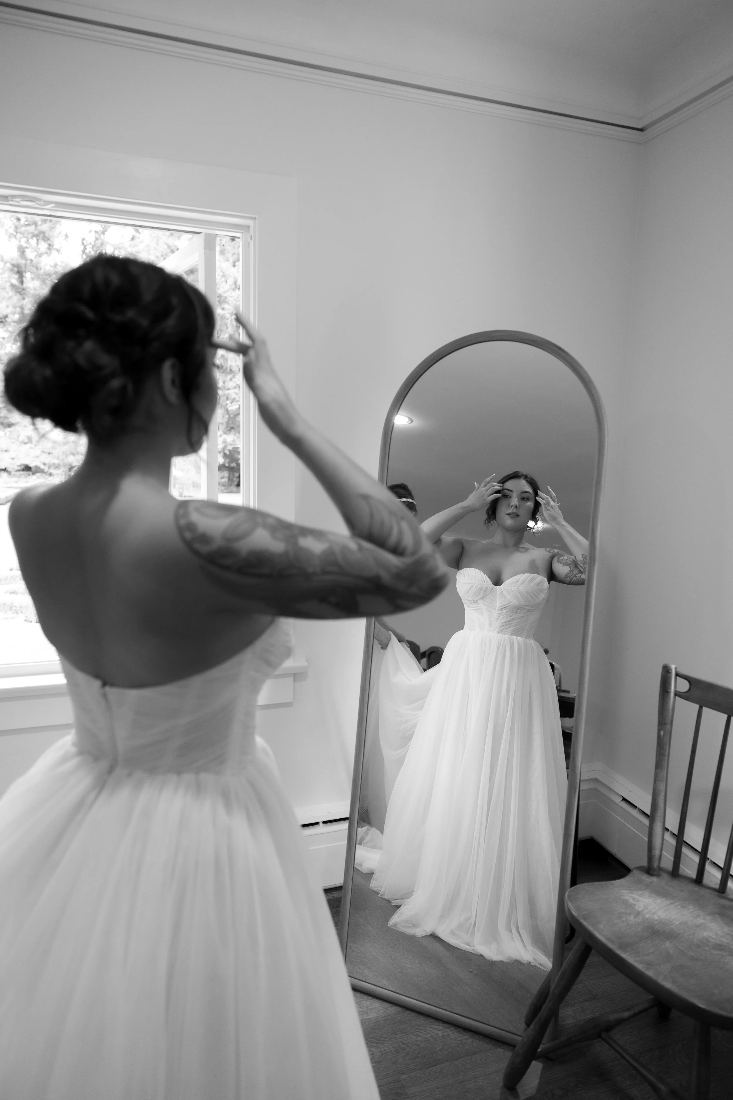 A woman in a wedding dress looks at herself in a full-length mirror while adjusting her hair.