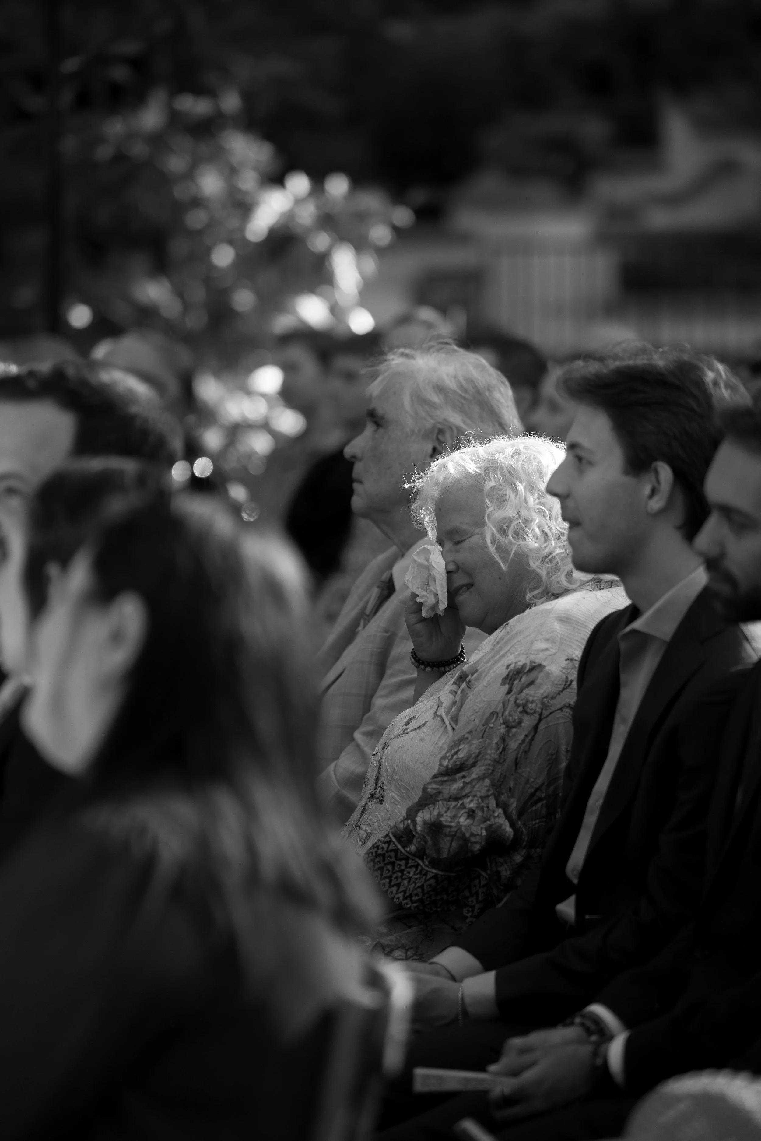 A black-and-white photo of people seated in rows at an indoor event, with one woman appearing to be crying and wiping her eye with a tissue.