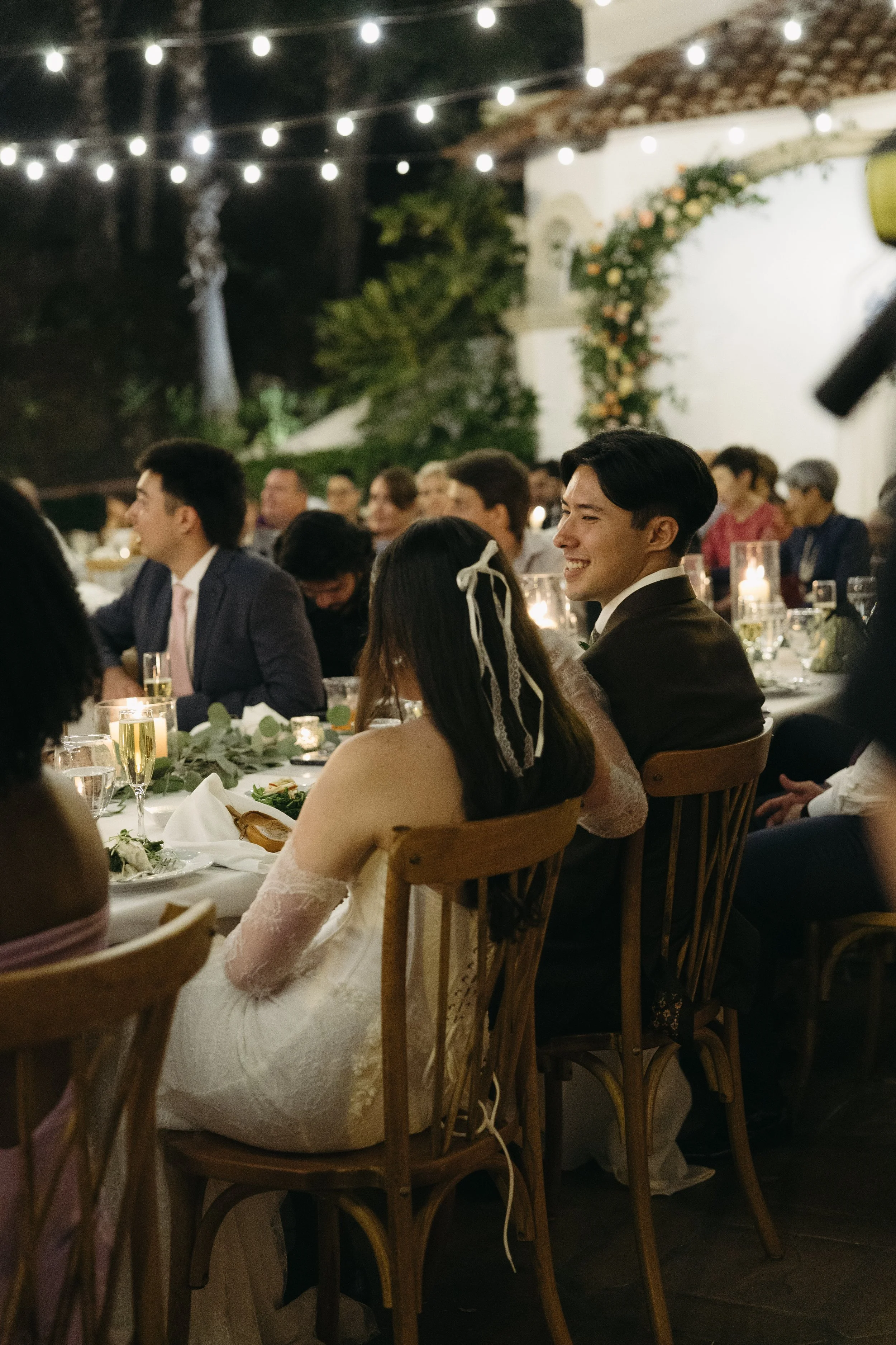 People attending a wedding reception outdoors at night, decorated with string lights and candles, seated at a long table.