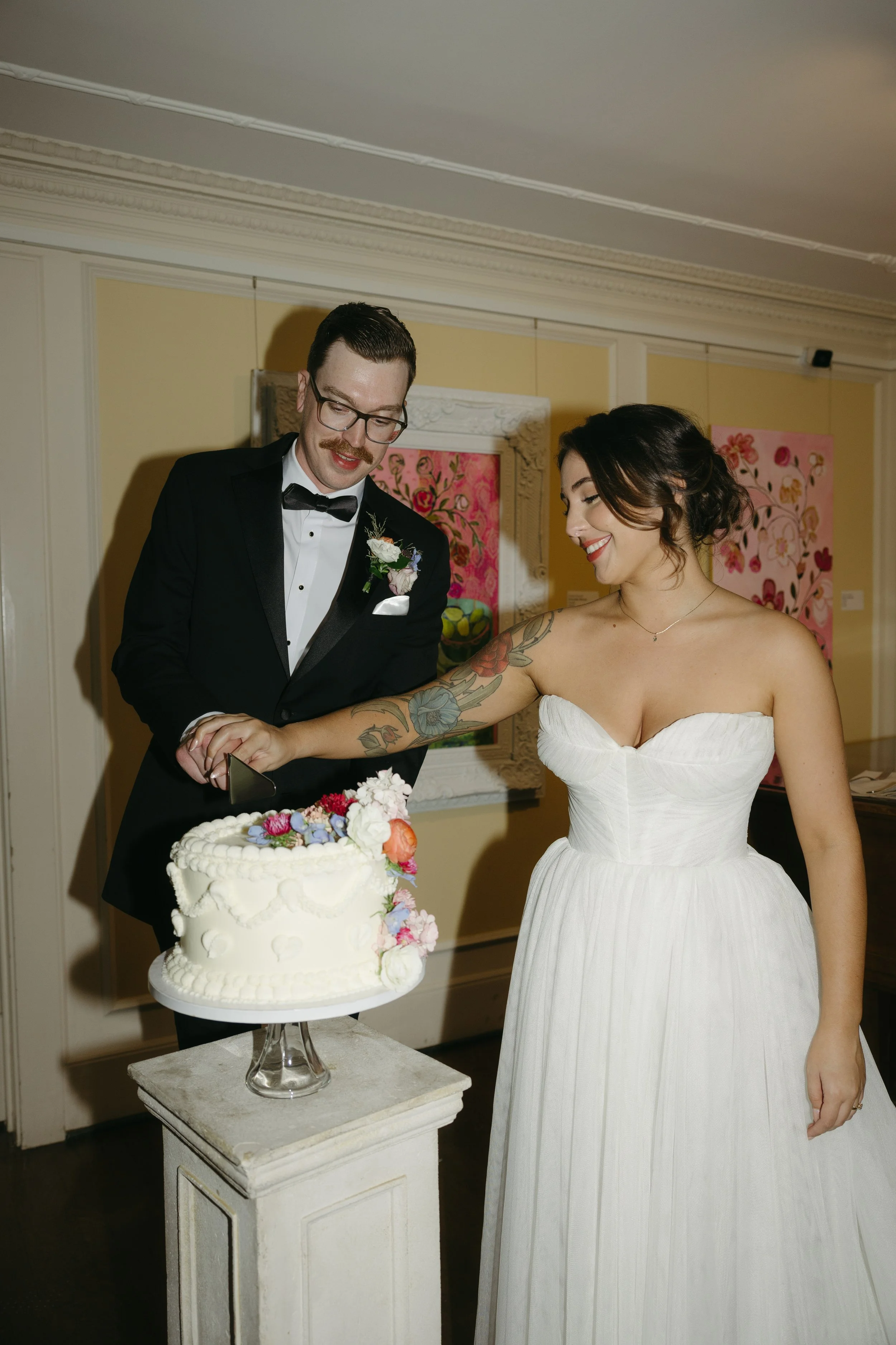 A bride and groom cutting their wedding cake, the bride in a white strapless gown with tattooed arm and the groom in a black tuxedo, in an indoor setting with artwork on the walls.