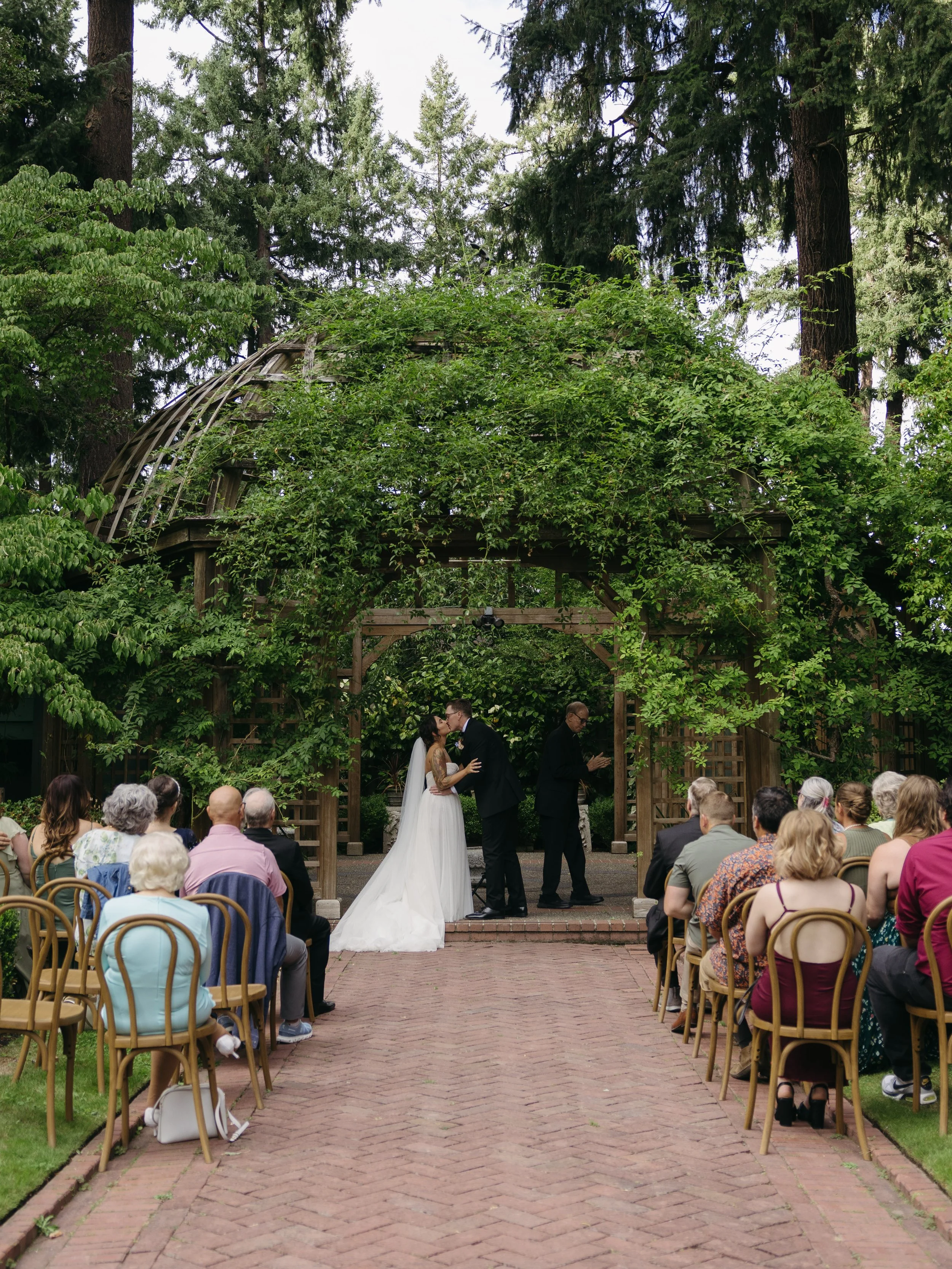 A wedding ceremony taking place outdoors in a lush, green garden with trees and foliage overhead. The bride and groom share a kiss at the altar, with guests seated on either side watching. An officiant stands nearby, reading from a book at a European