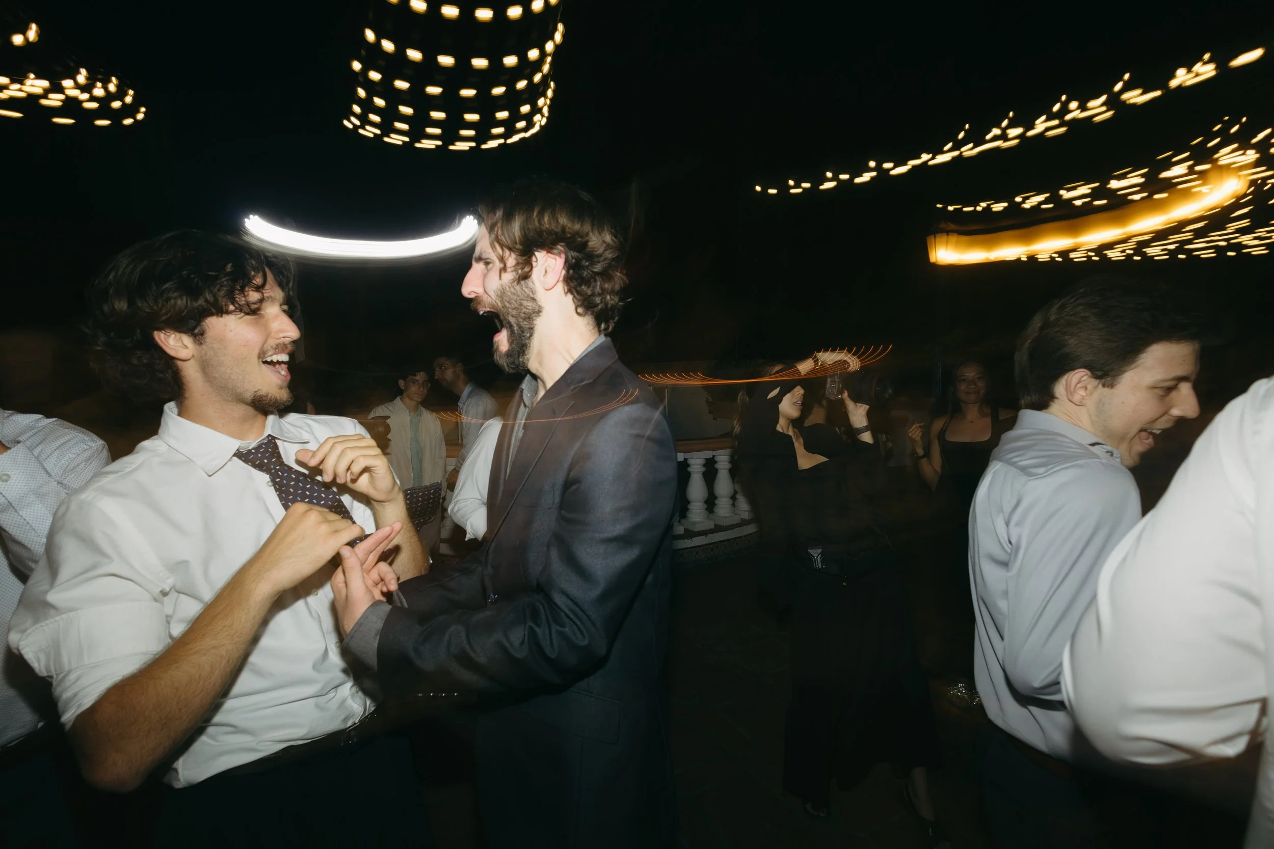 Two men partying at a nightclub, dancing and smiling with people in the background. The scene is lively with light streaks and dim lighting.