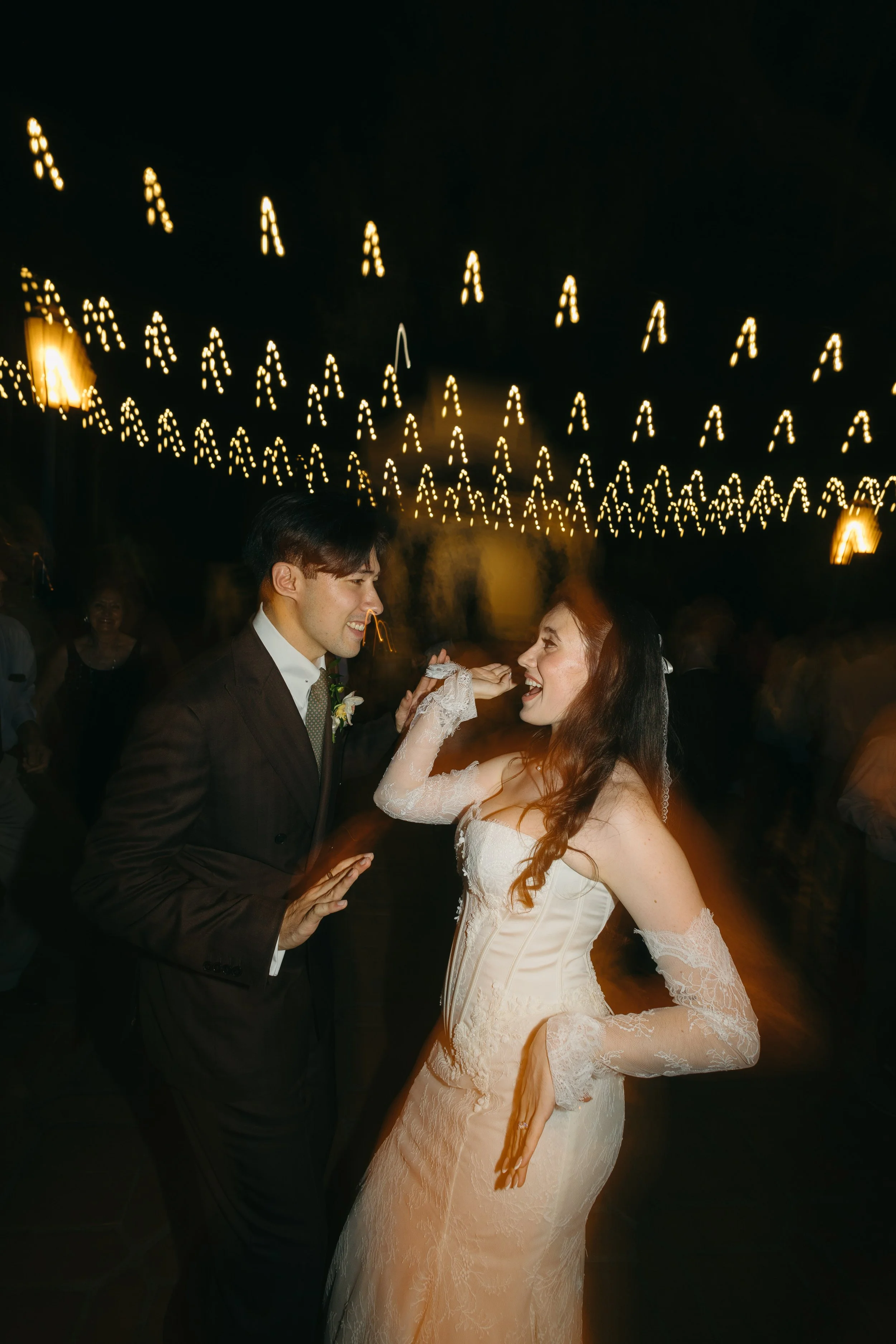 A bride and groom dancing at their wedding reception with string lights overhead at night.