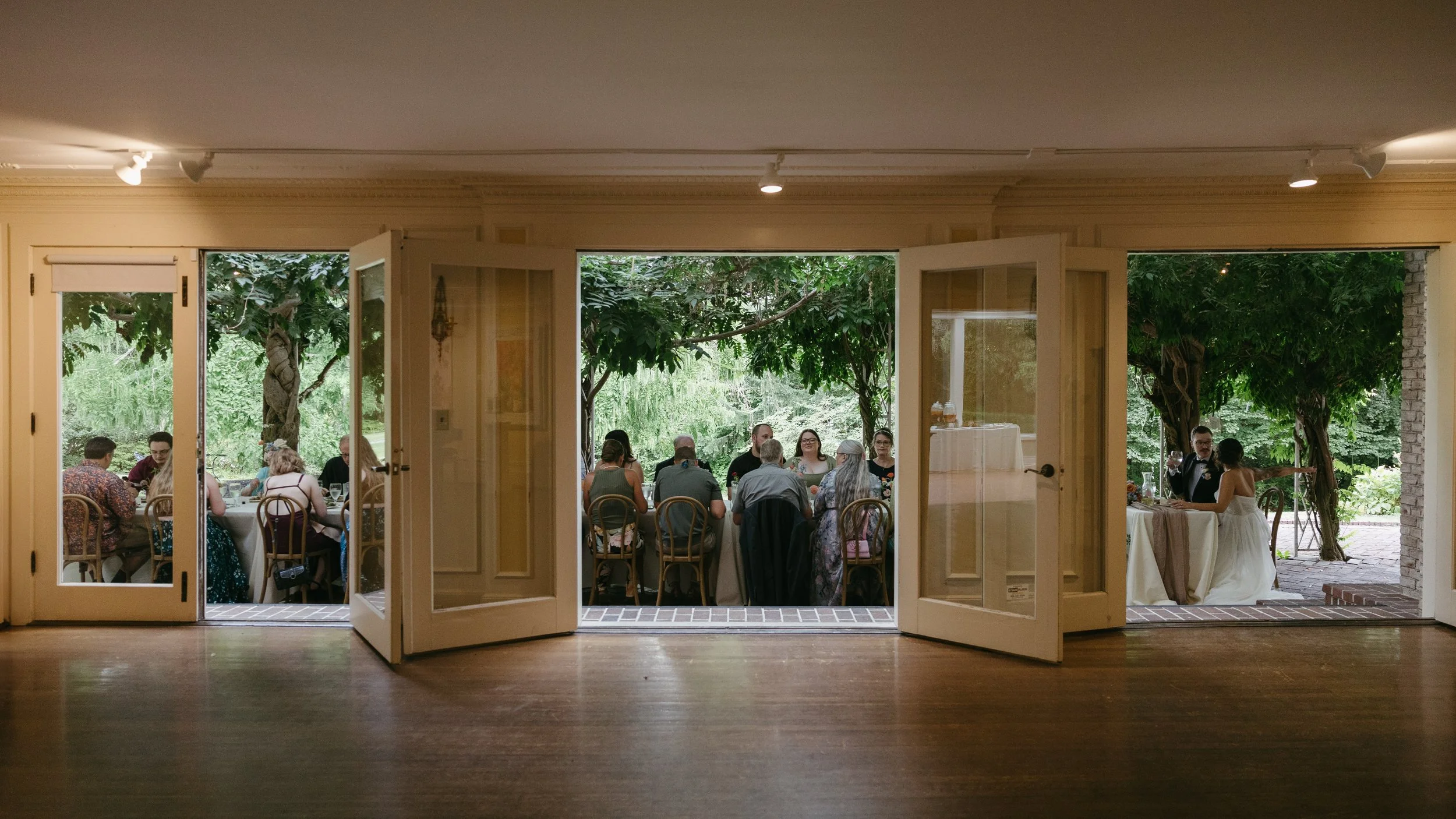 View of a wedding reception outdoors through open French doors, with guests seated at tables under trees.