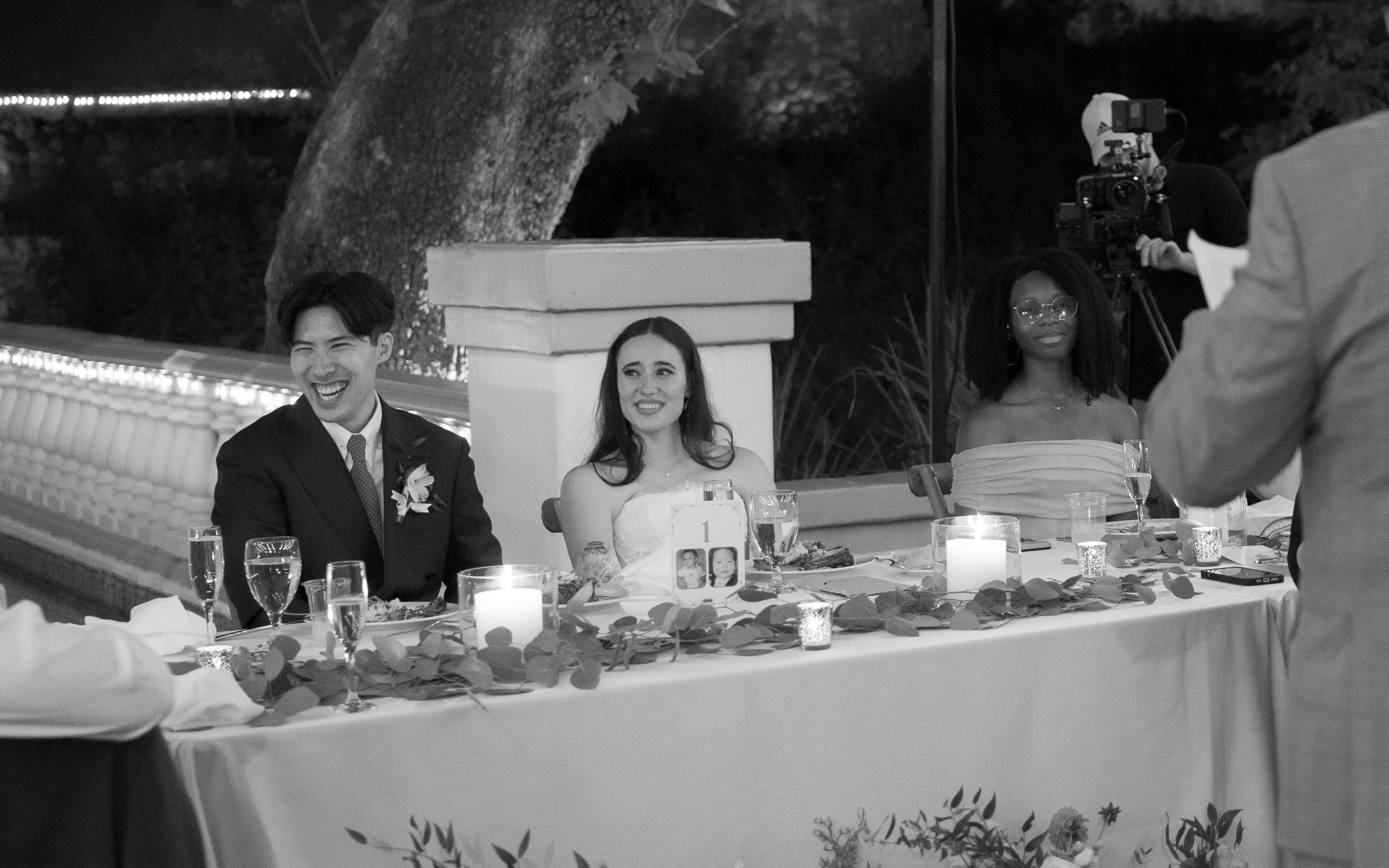 A wedding reception scene with three women sitting at a decorated table. The woman in the middle is the bride, wearing a strapless dress, and the woman on the right is wearing glasses and an off-the-shoulder dress. A man on the left is wearing a suit