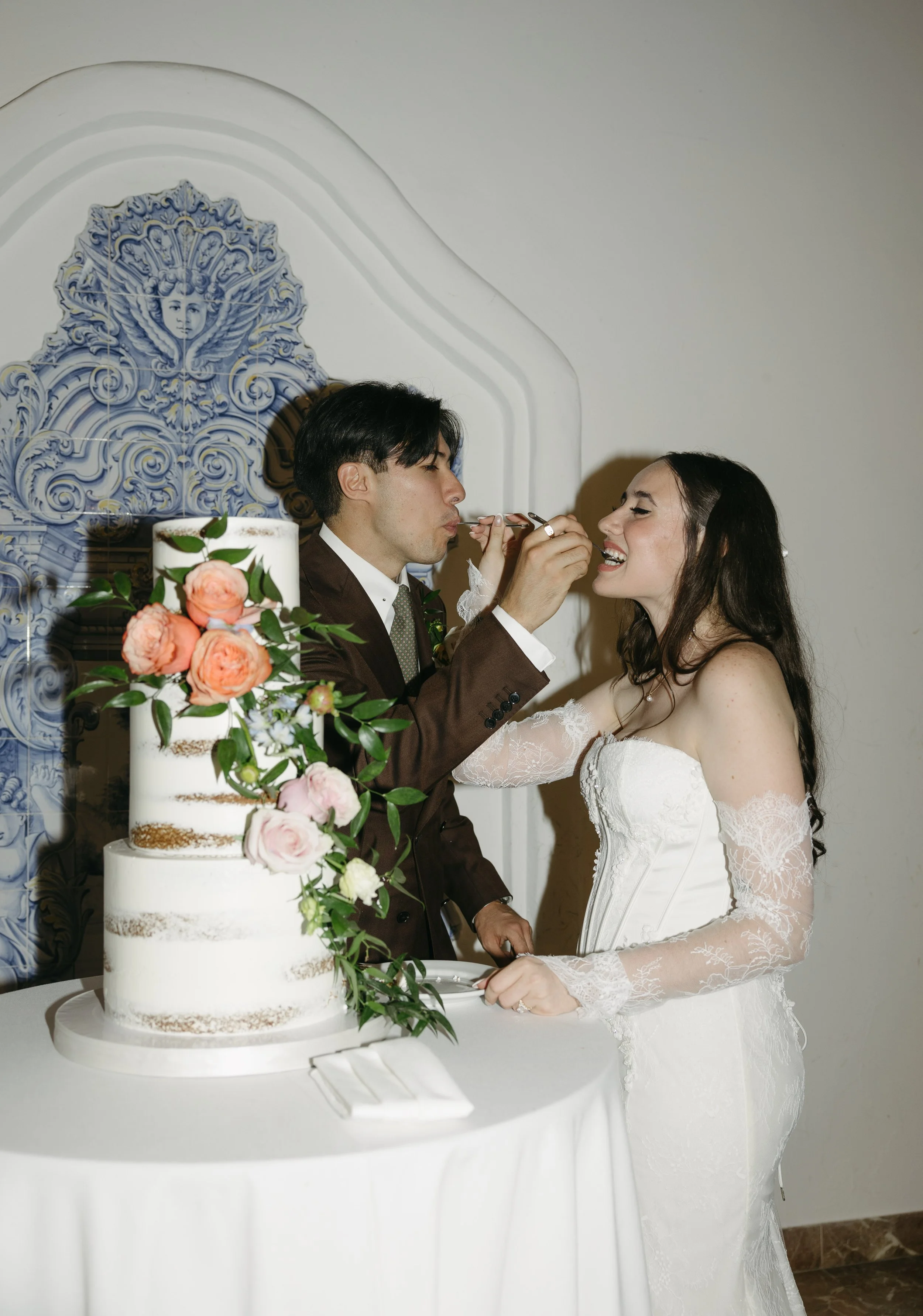 A bride and groom are feeding each other cake at their wedding celebration, with a multi-tiered wedding cake decorated with pink and white roses in the foreground.