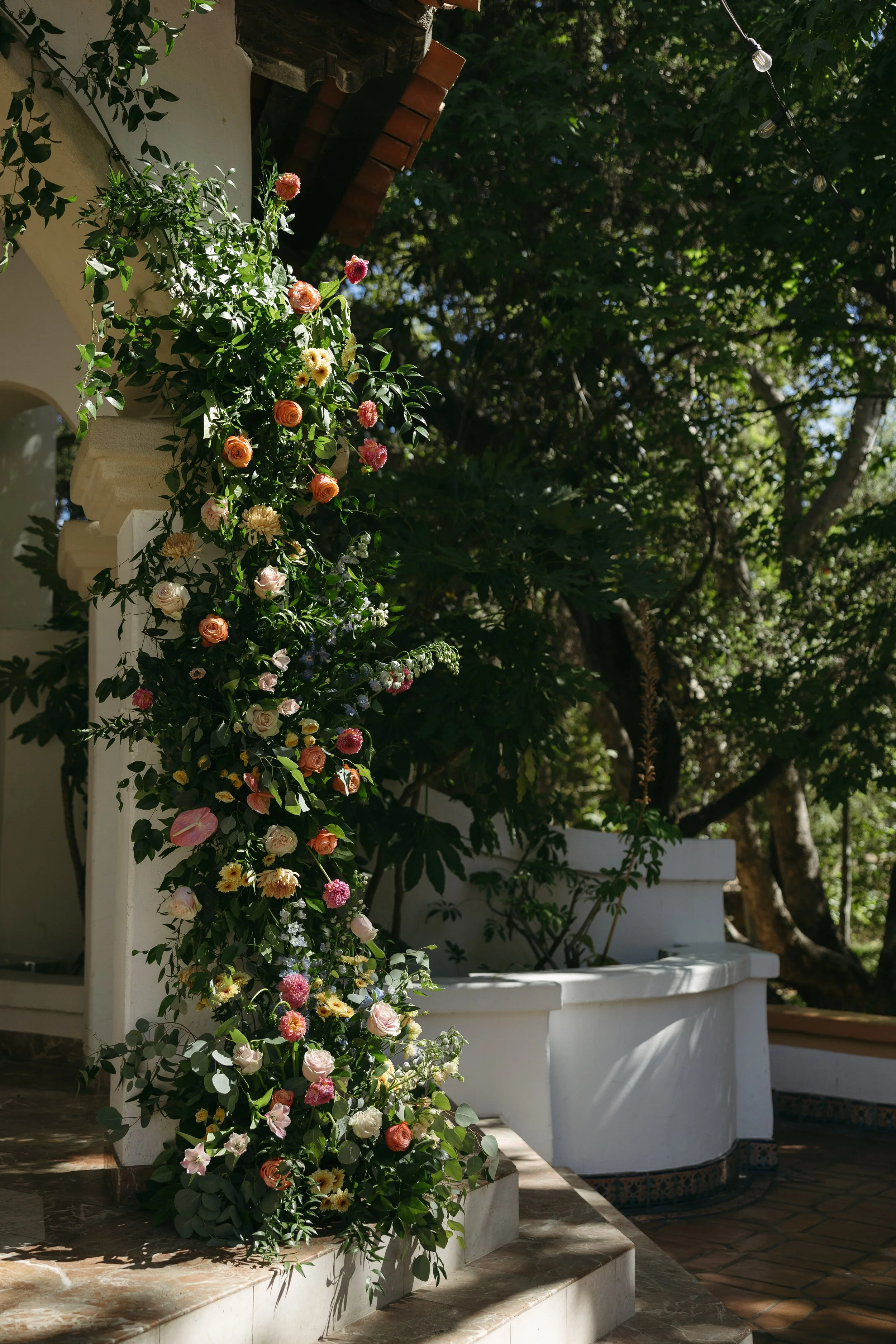 A tall floral arrangement of various colorful flowers, including roses and dahlias, cascading down a white wall next to a staircase outside a house, with lush green trees in the background.