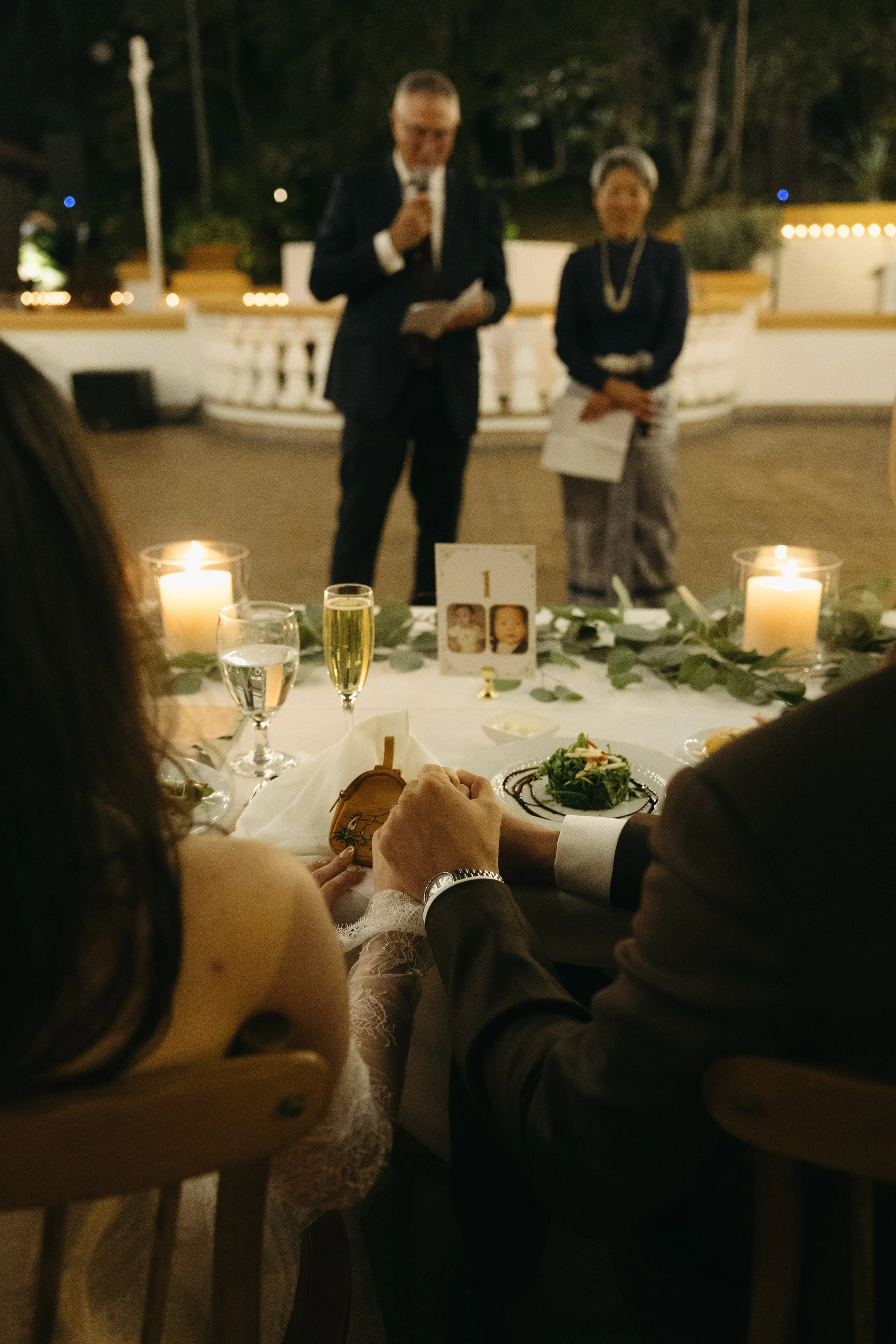 A wedding reception with a table set with candles, drinks, and a salad, where a couple is holding hands, and a man and woman are giving speeches in the background.