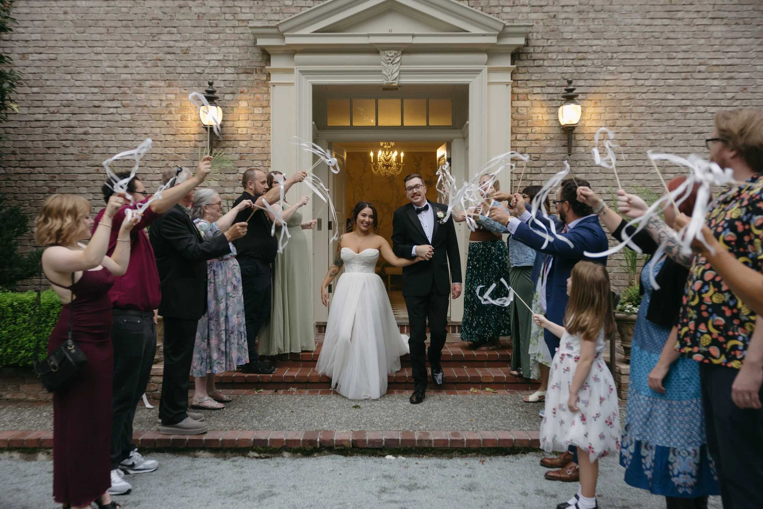 A newlywed couple walking out of a building, holding hands, while wedding guests celebrate their sendoff with ribbons outside on a brick staircase at a European inspired Lakewold Gardens wedding in Seattle