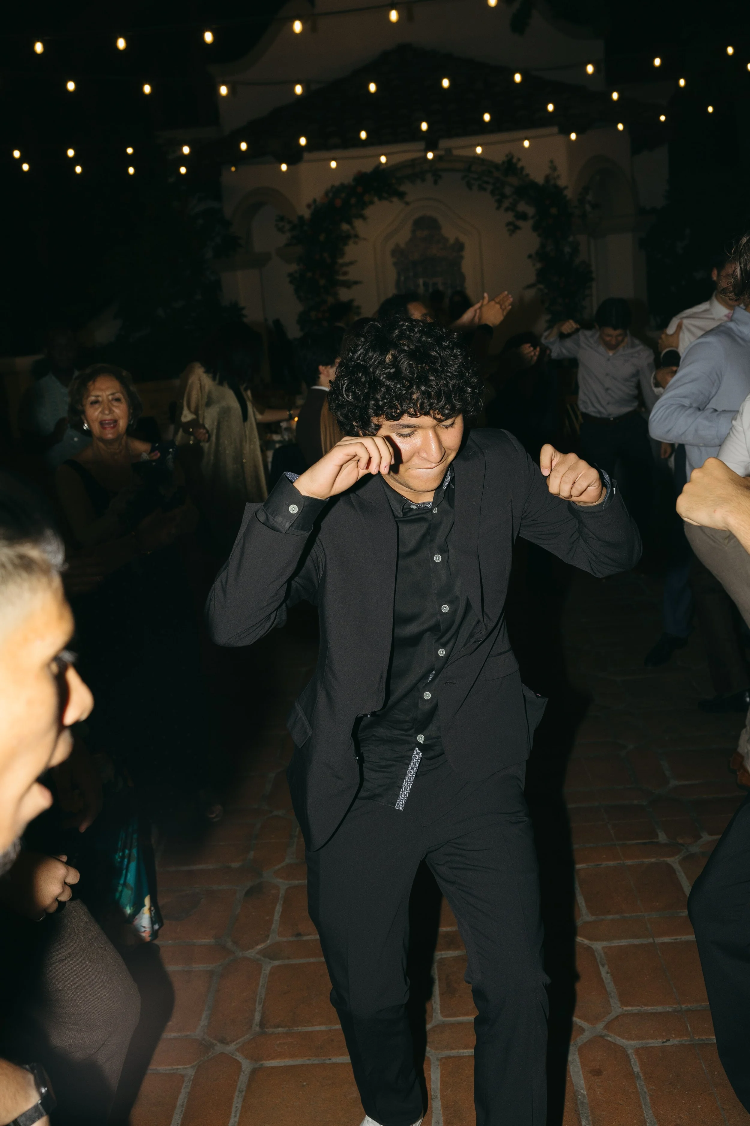 A young man in a black suit is dancing with a joyful expression at a celebration, surrounded by other people dancing and enjoying the event under string lights.
